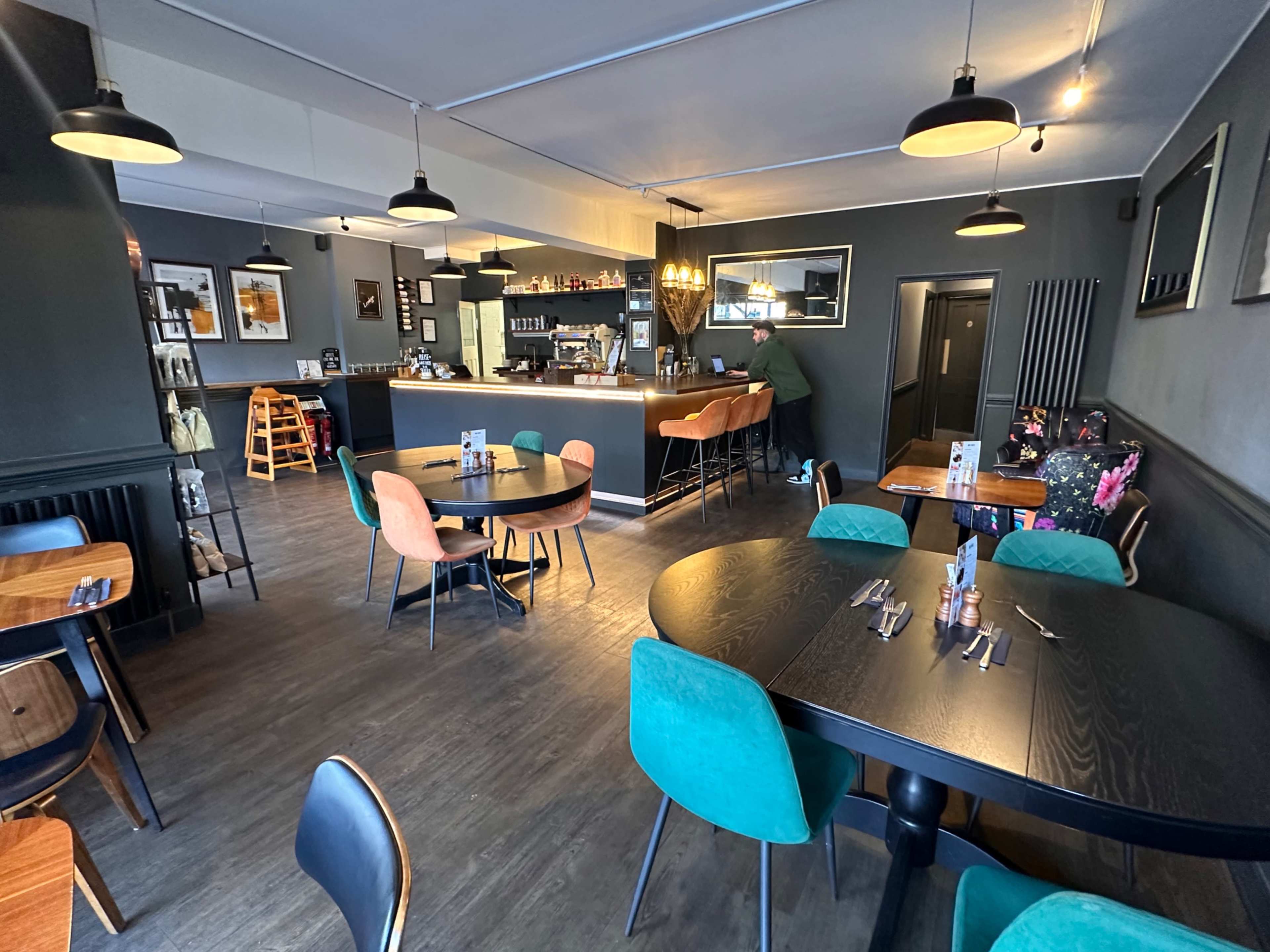 The image shows a modern restaurant interior with wooden tables, colorful chairs, and a bar area in the background.