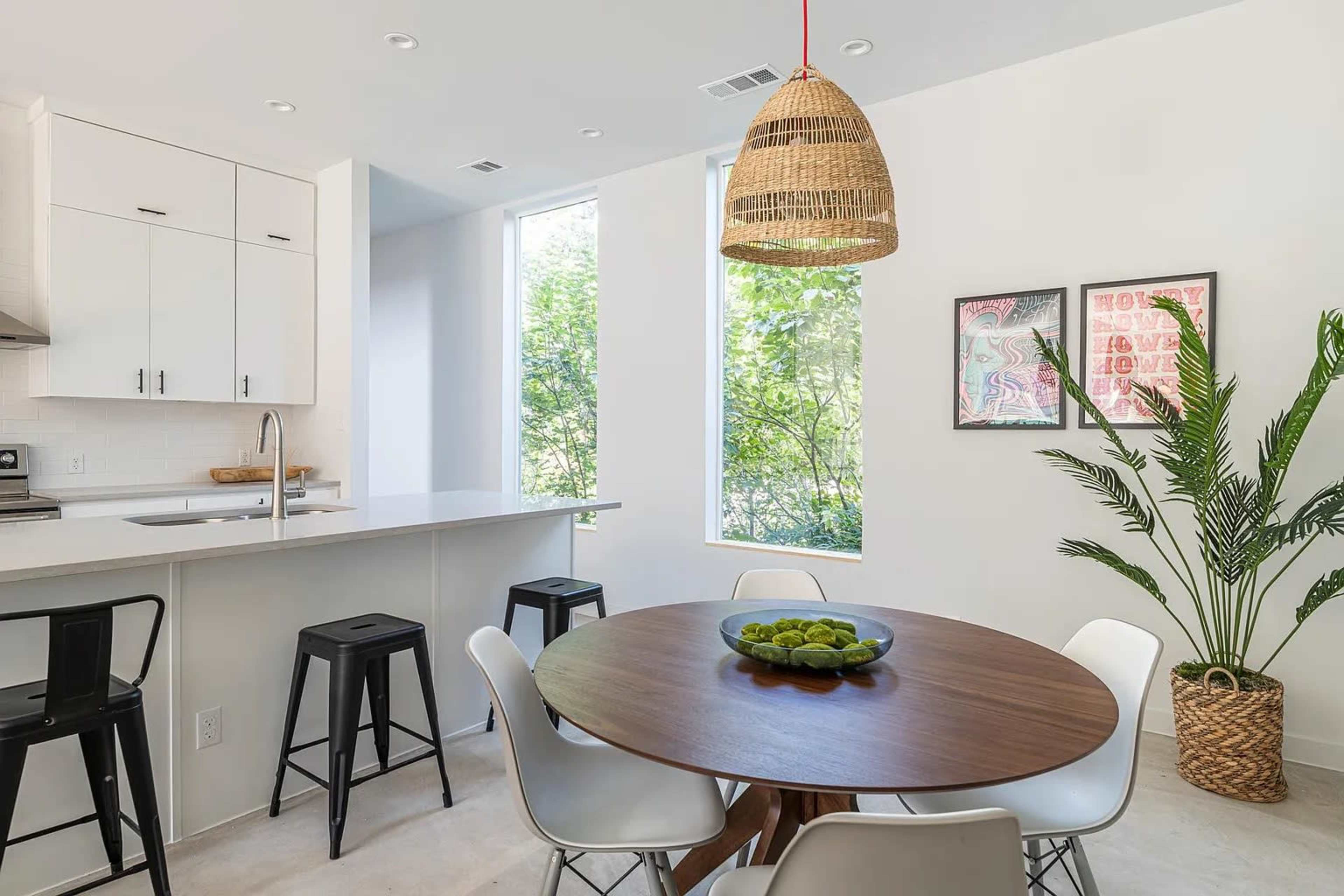 A dining area features a round wooden table surrounded by white chairs and black stools, with large windows providing natural light and a potted plant nearby.