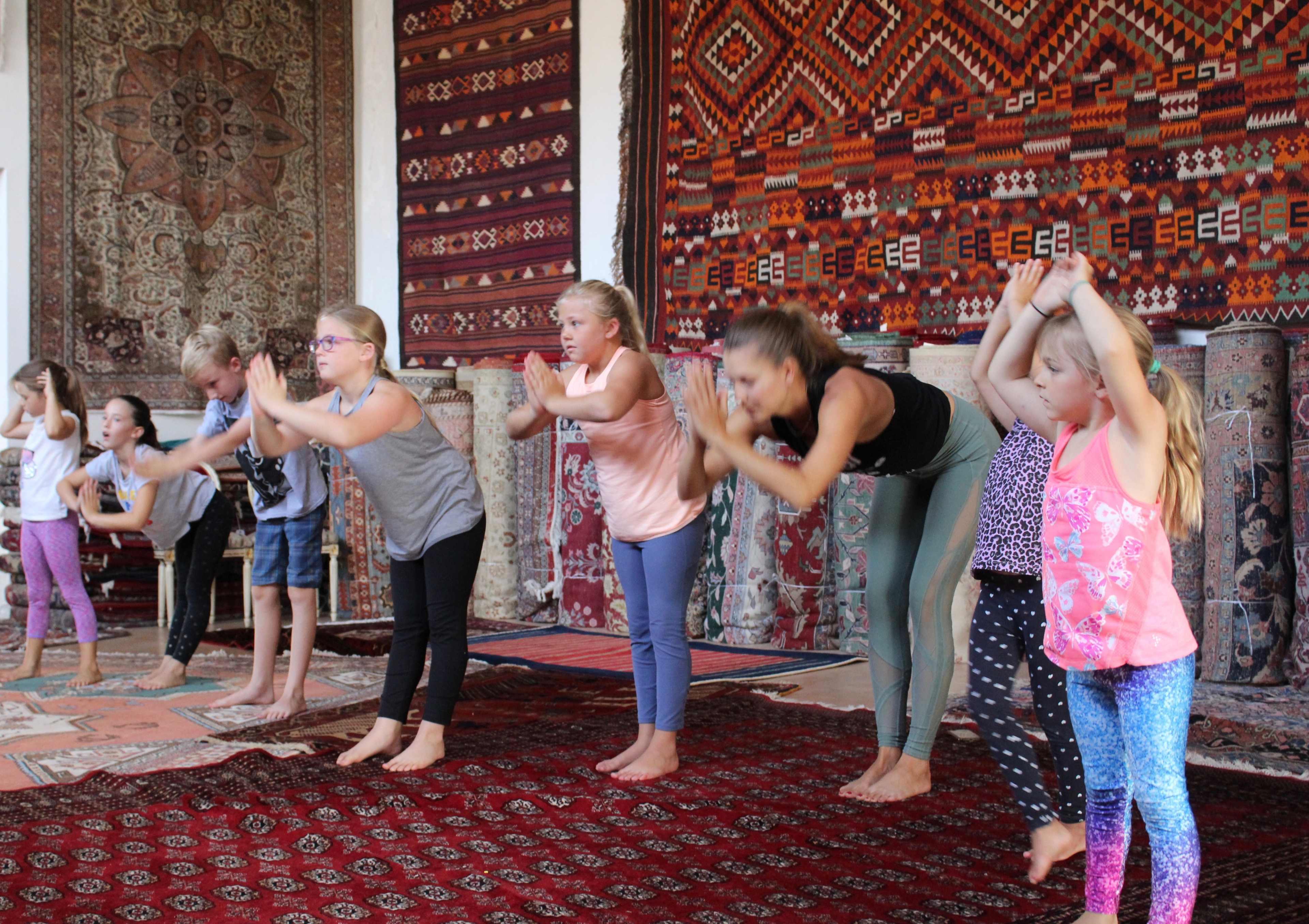 A group of children and one adult practice yoga poses in a decorated room with patterned carpets and tapestries.