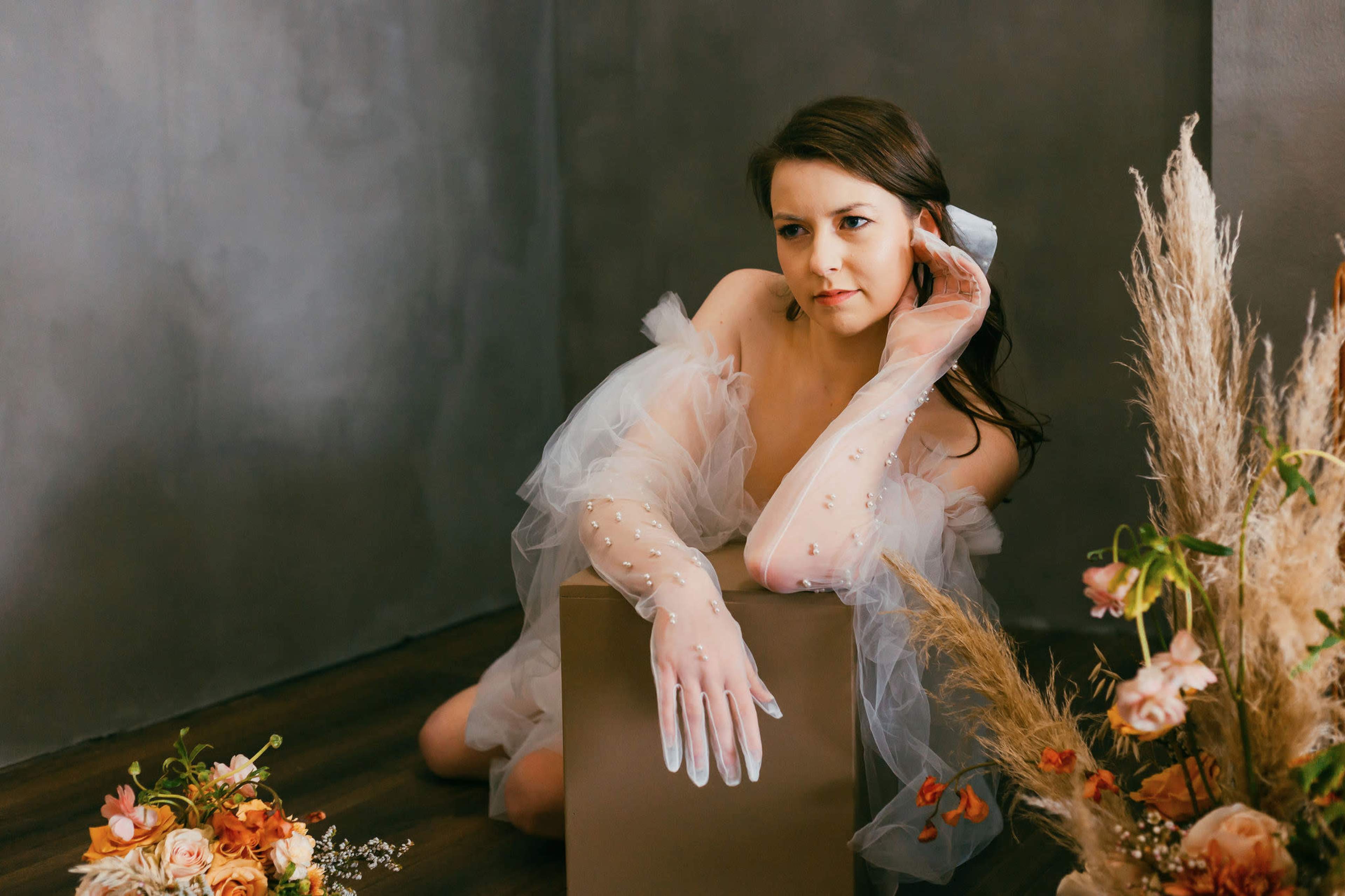 A woman in a sheer, ruffled outfit poses gracefully beside a beige box, surrounded by floral arrangements on a wooden floor against a gray backdrop.