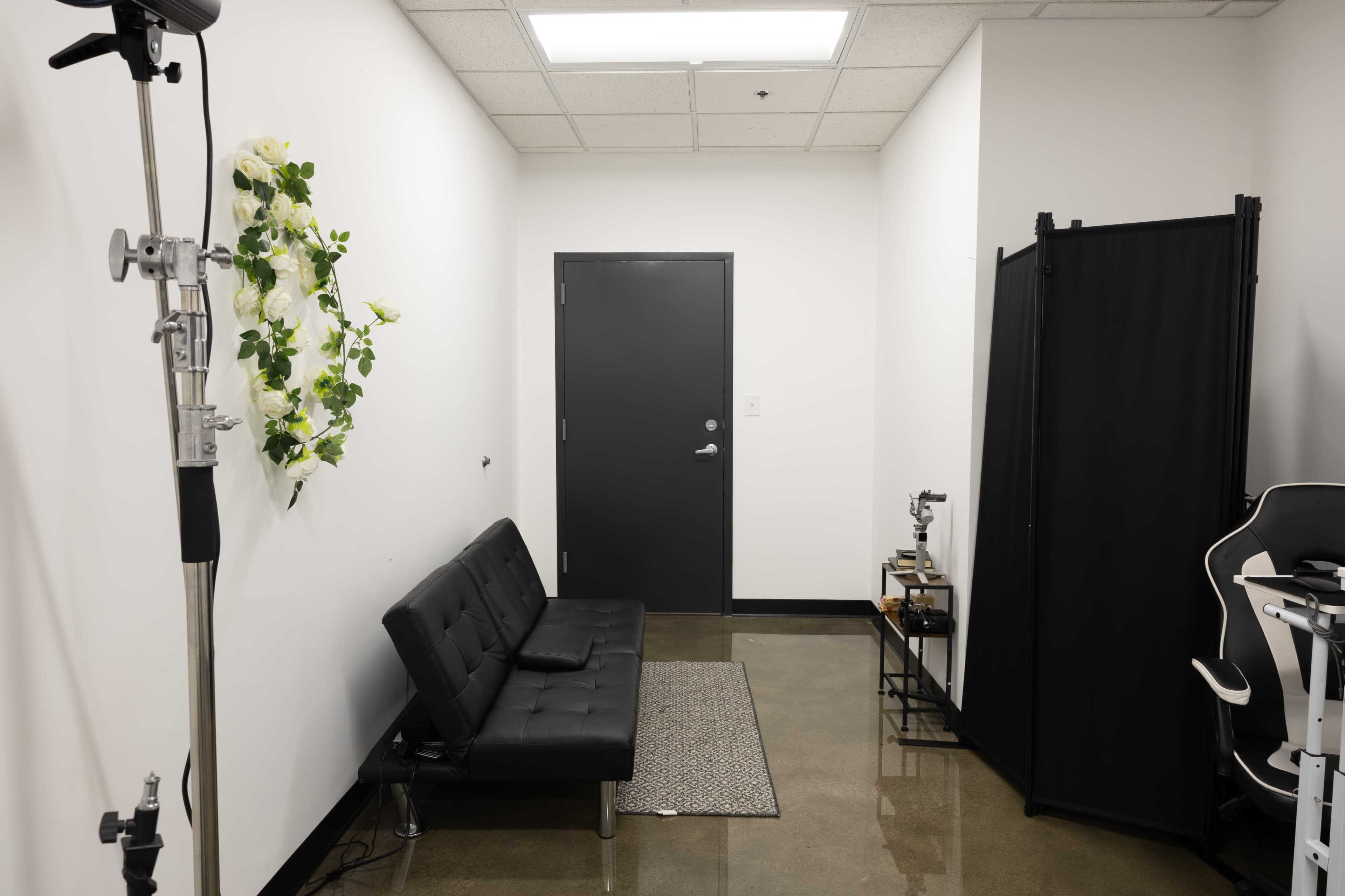 The image shows a small waiting area with a black sofa, a decorative floral arrangement on the wall, and a door leading to another room.