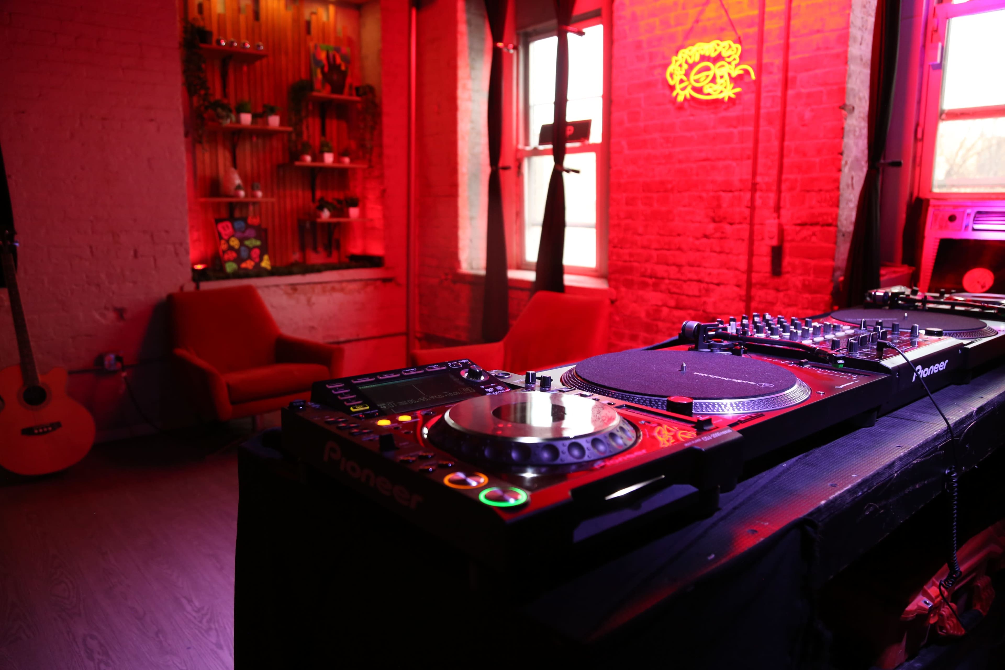 A DJ setup with turntables and a mixer is positioned in a dimly lit room featuring red lighting and a decorative wall.