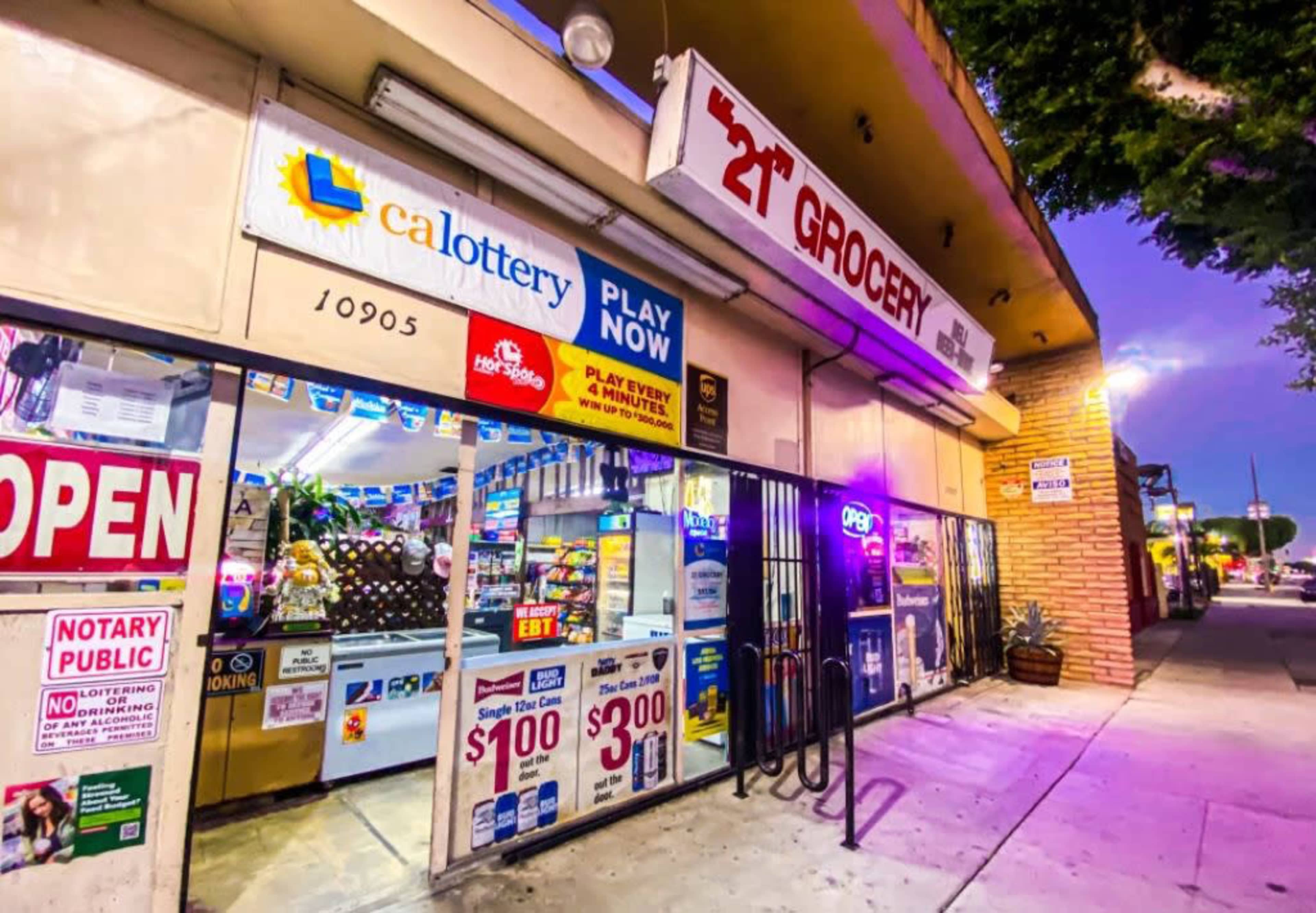 A grocery store with bright signage and a display of lotto tickets is open for business on a city street at dusk.
