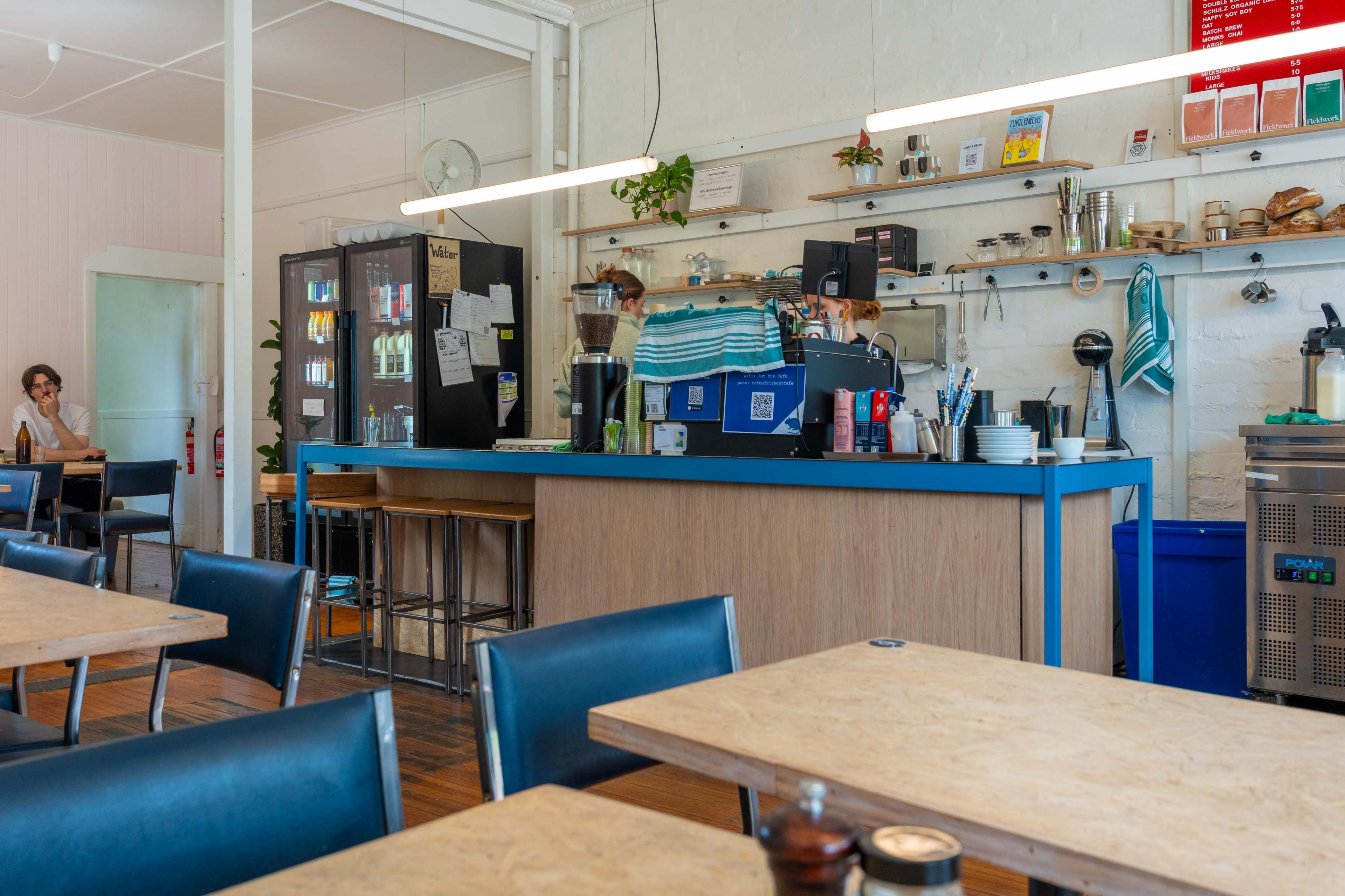 The image shows a modern cafe interior featuring a wooden counter with a coffee machine, shelves lined with utensils, and tables set for customers.