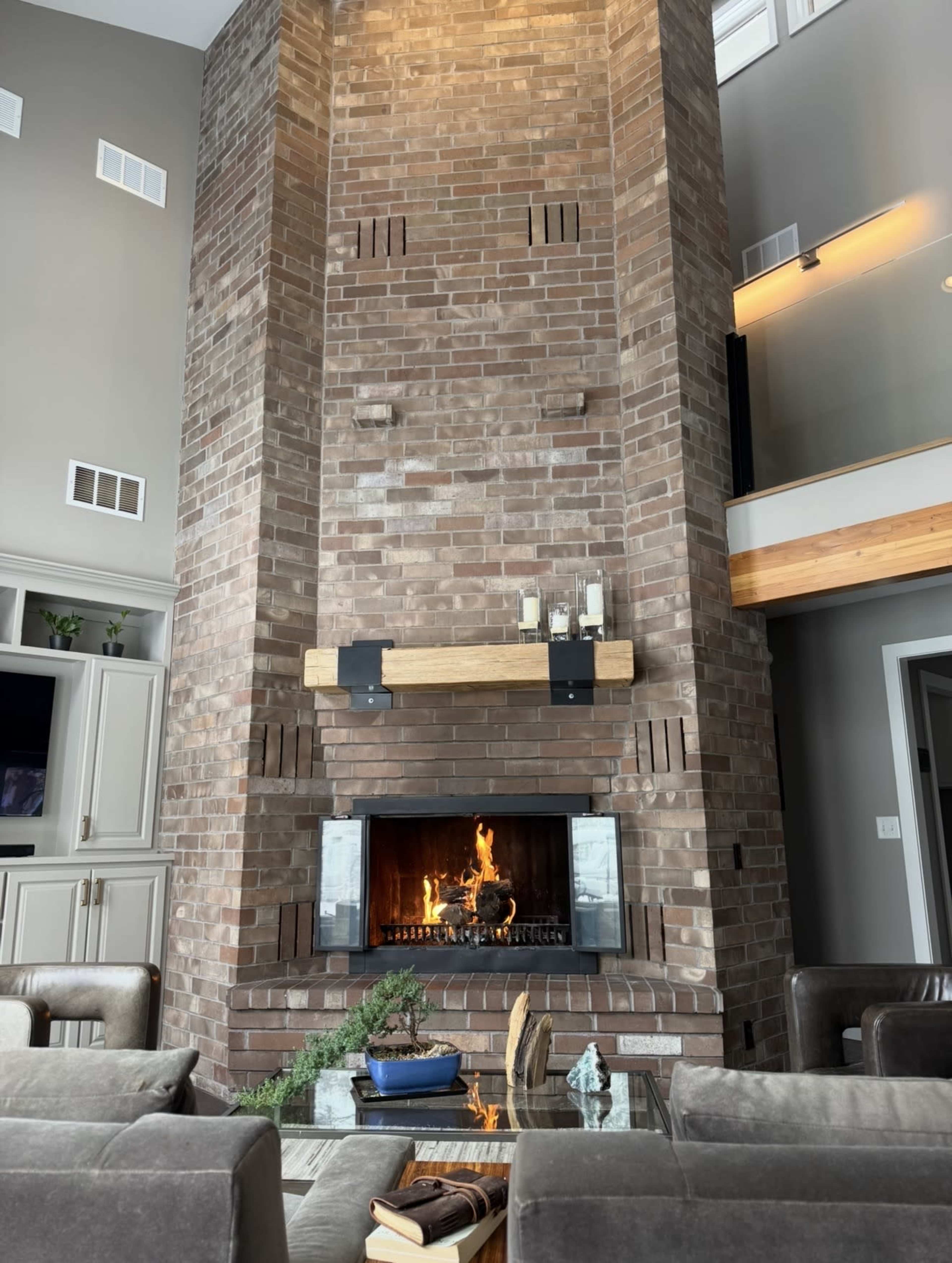 The image shows a modern living room featuring a tall brick fireplace with a fire burning, surrounded by comfortable seating and a wooden beam mantel.