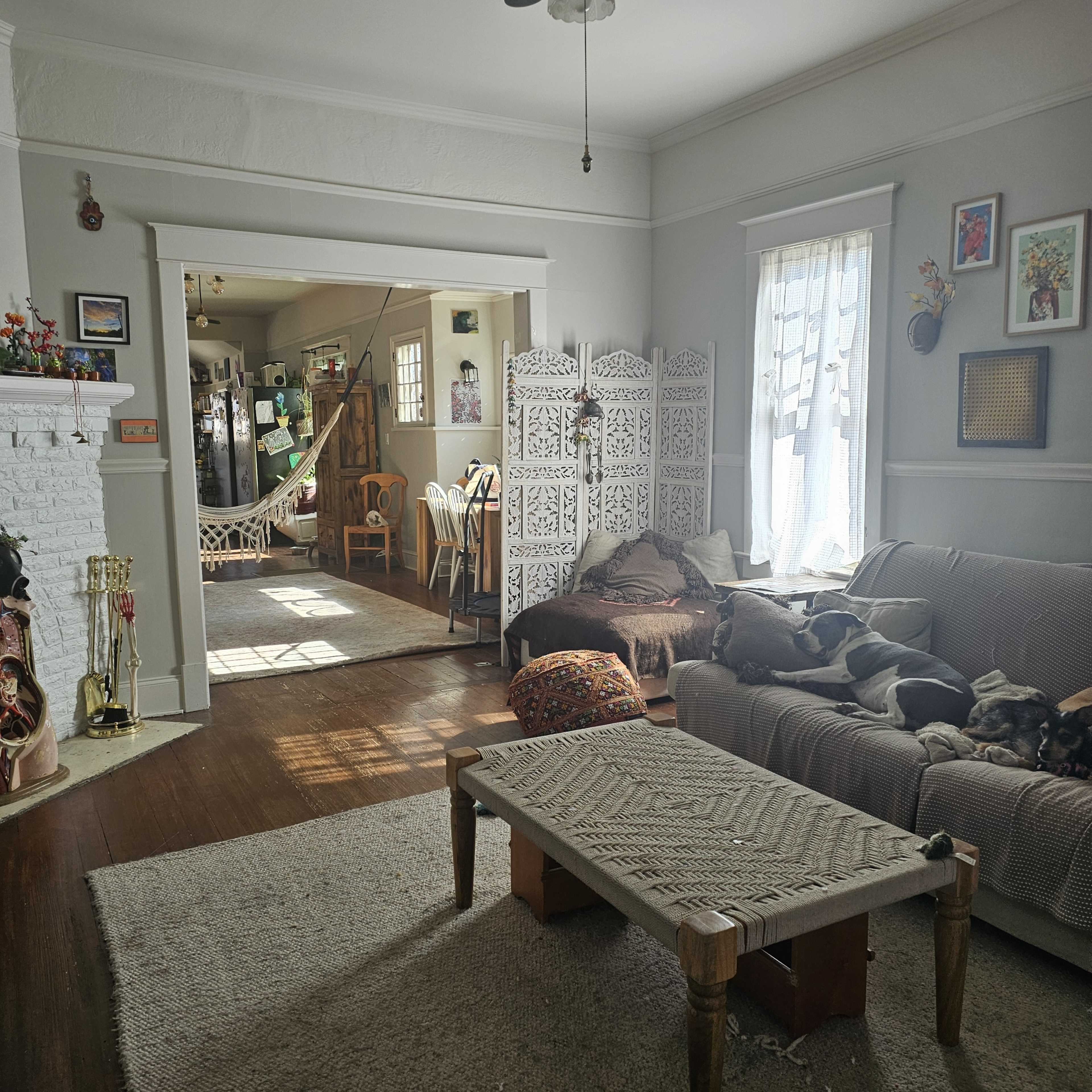 The image shows a cozy living room with a sofa, a low wooden table, a patterned rug, and a partition, leading to a dining area with chairs and a visible kitchen in the background.
