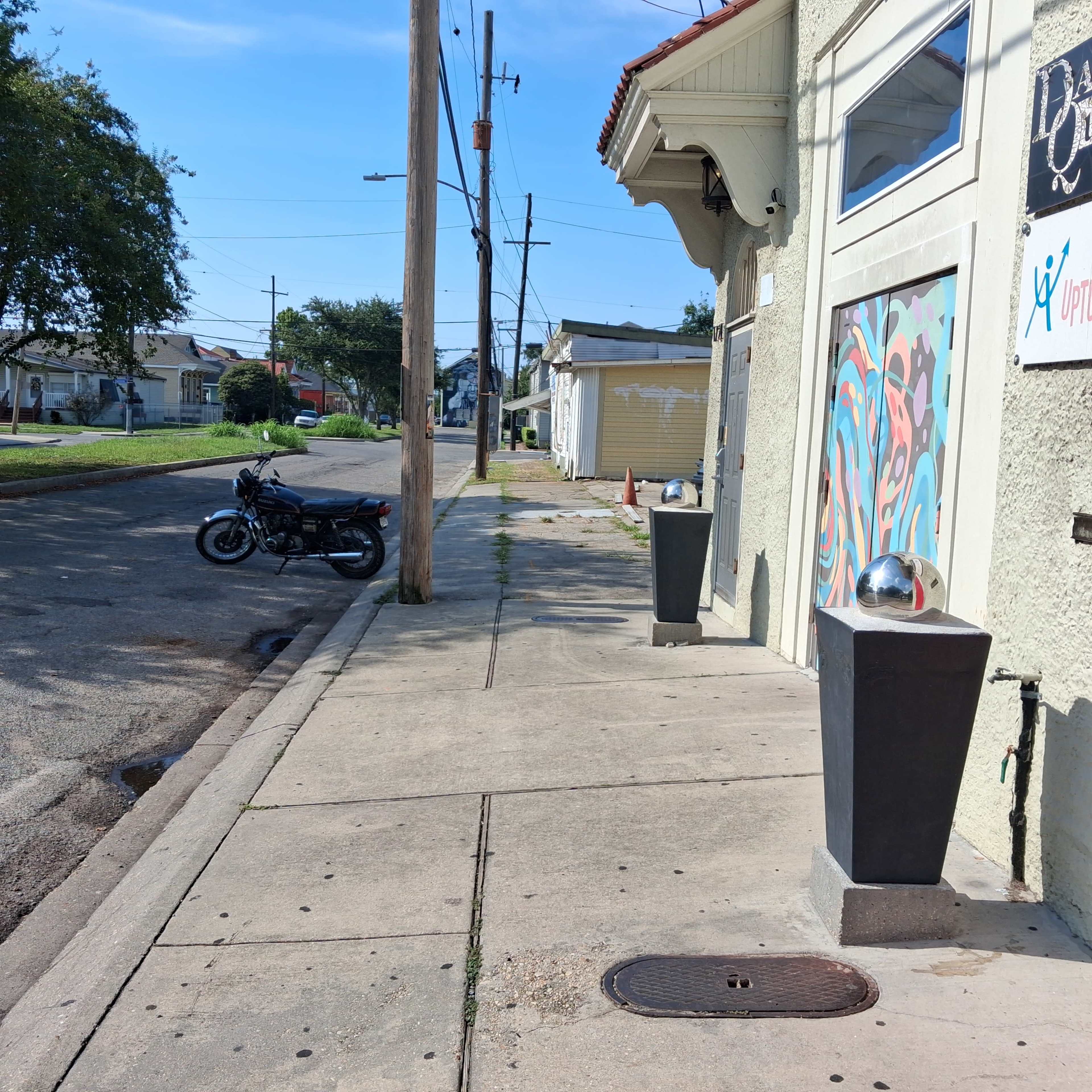 The image shows a sidewalk next to a street, featuring a parked motorcycle and a building with a colorful mural on its side.