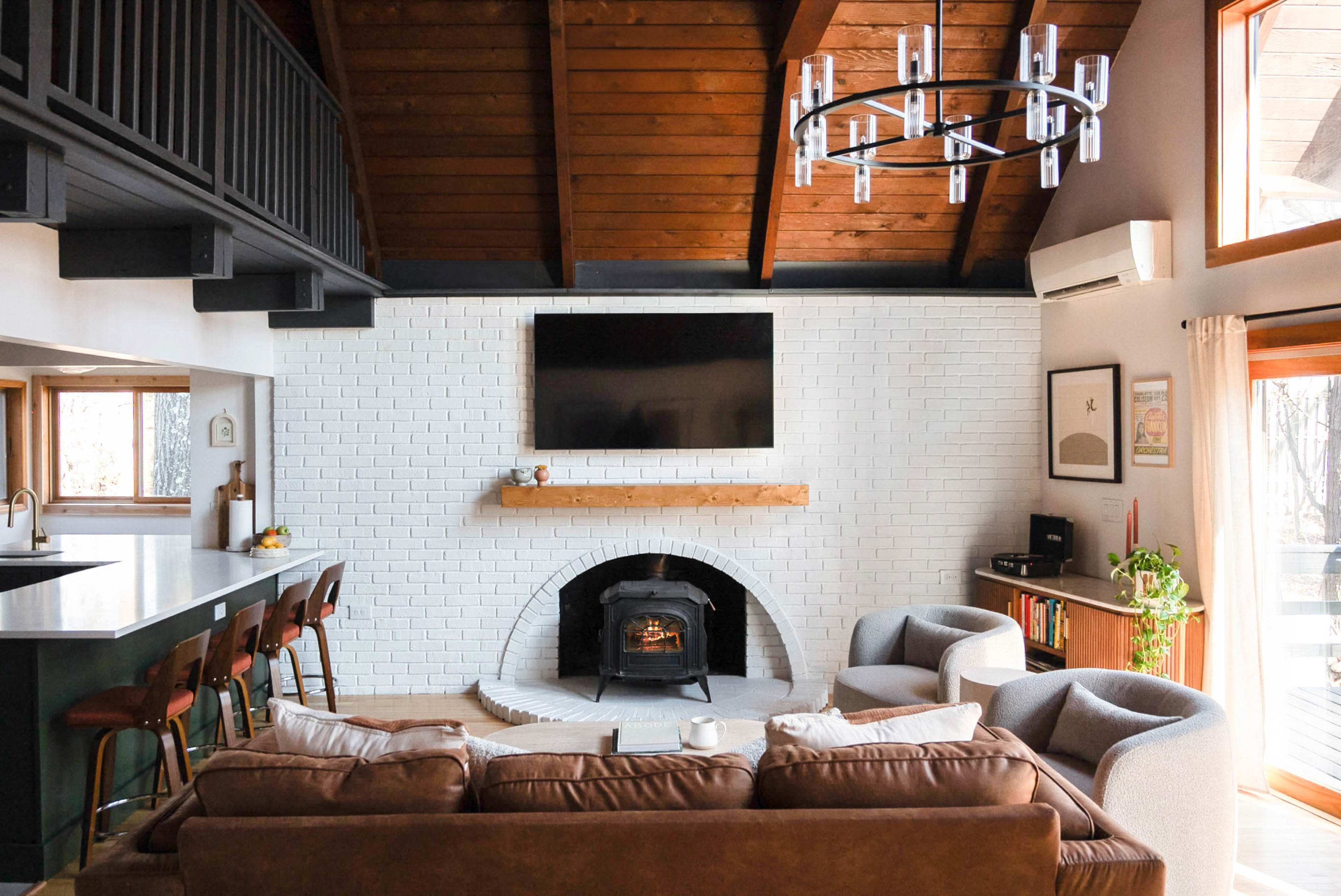 The living room features a white brick fireplace, a large flat-screen TV mounted above a wooden mantel, and a seating area with a brown sofa and gray armchairs.