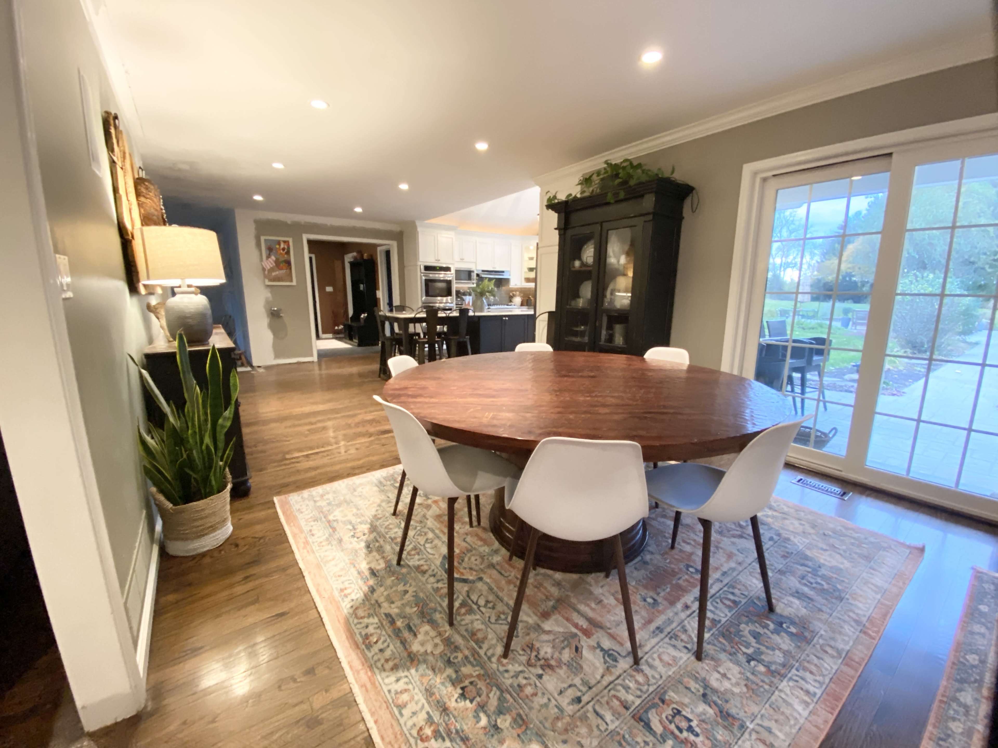 A round wooden dining table surrounded by white chairs is set in a well-lit room with hardwood floors and large sliding glass doors leading to a patio.