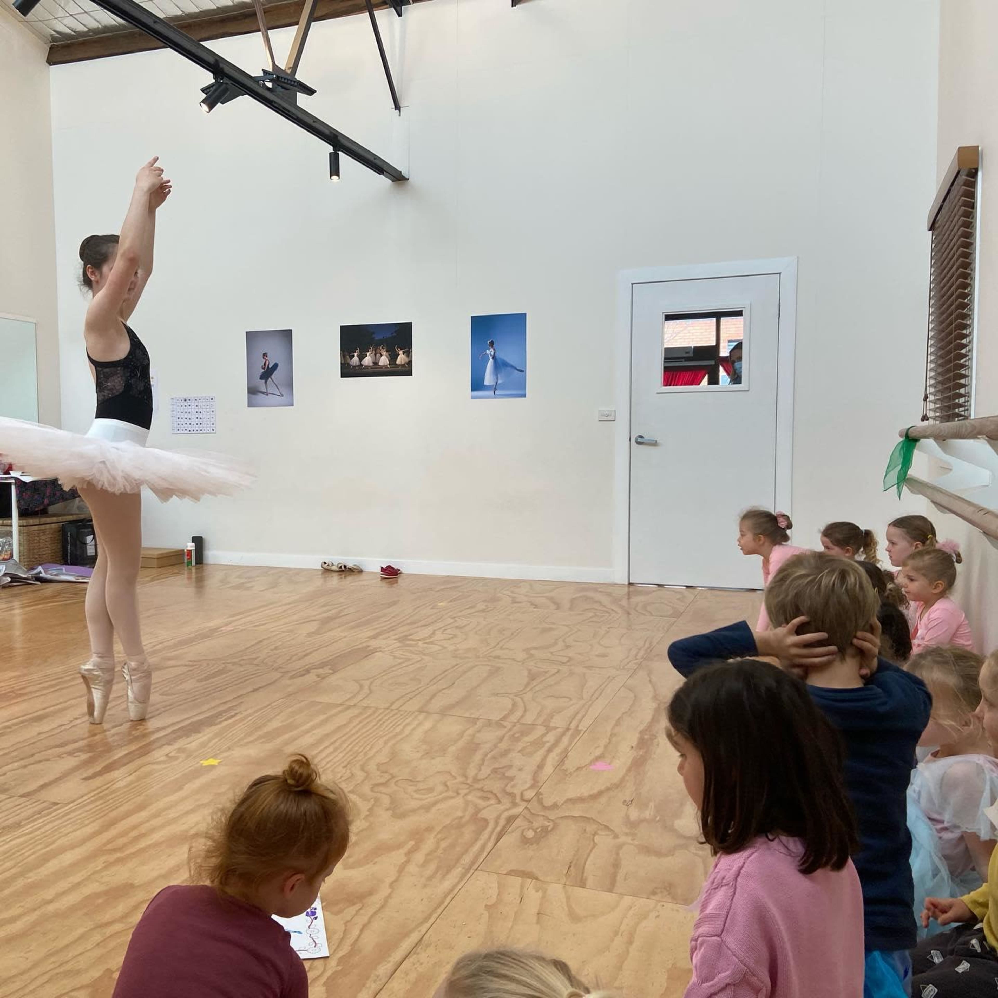 A ballet dancer performs on pointe in a dance studio while a group of attentive children watches from the sidelines.