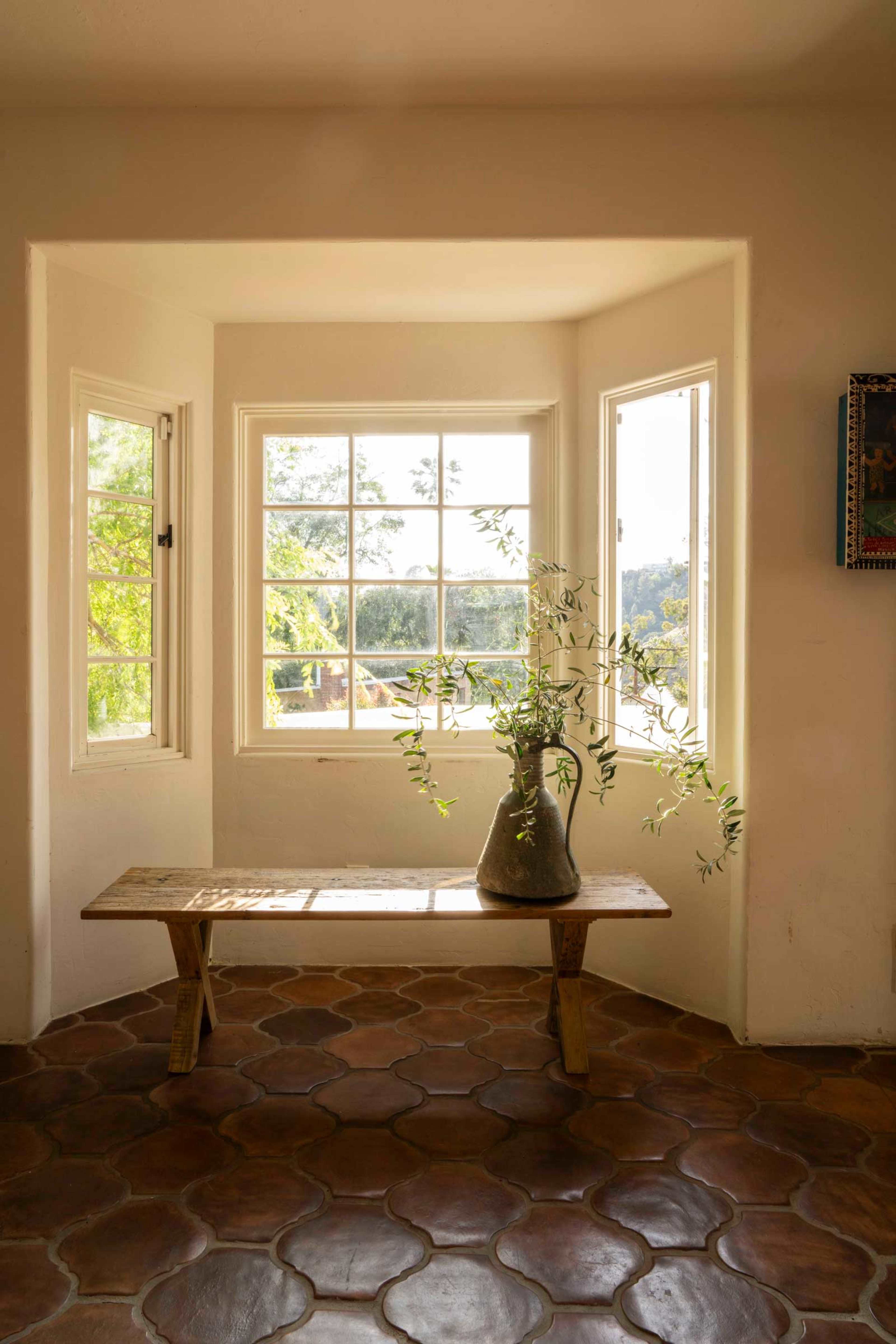 A wooden bench sits beneath a trio of windows, with a vase of greenery on top, in a sunlit corner of a room with tiled flooring.