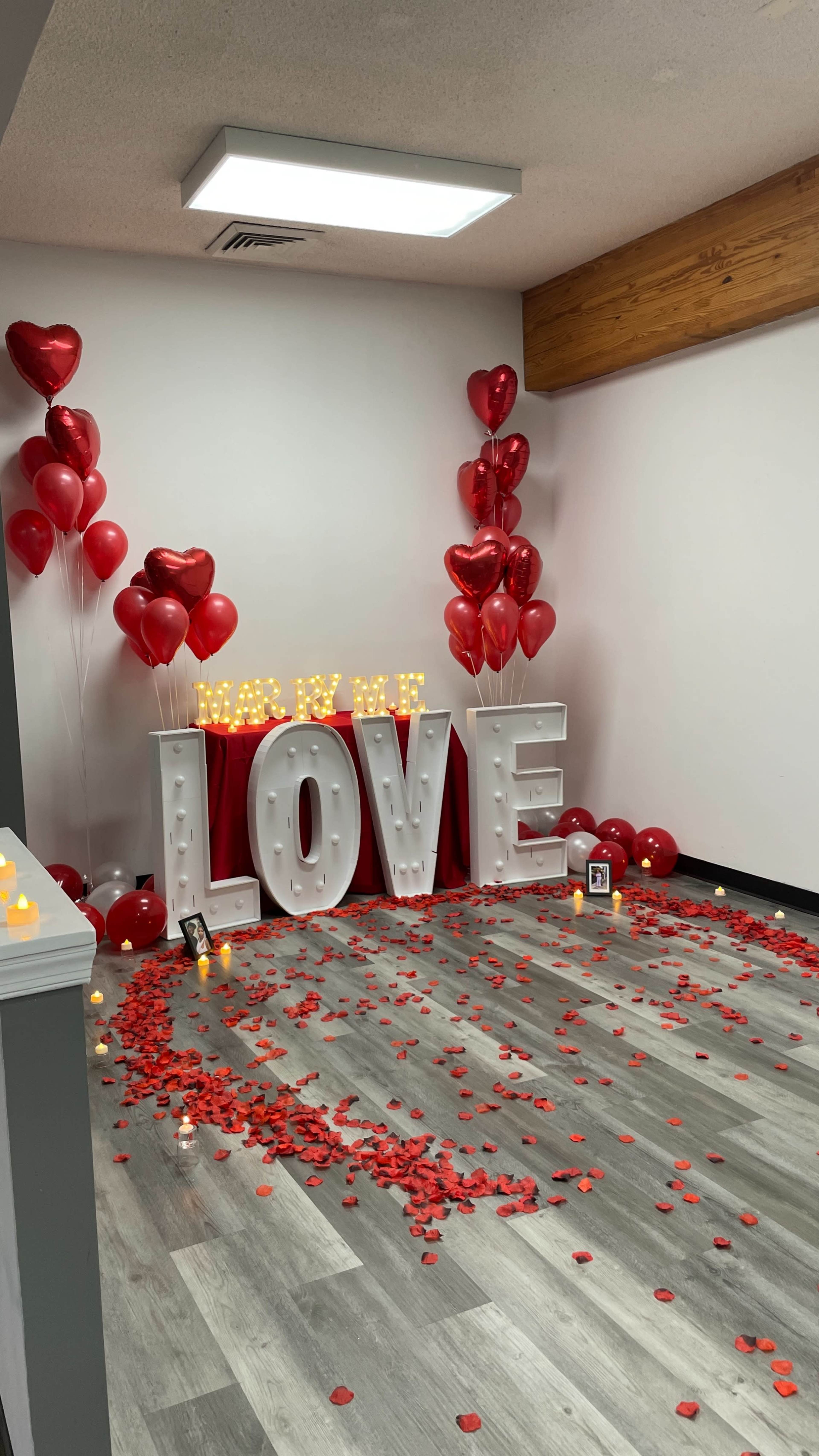 The scene features large illuminated letters spelling "LOVE," surrounded by red balloons, a red table, and rose petals on the floor, creating a romantic setting.