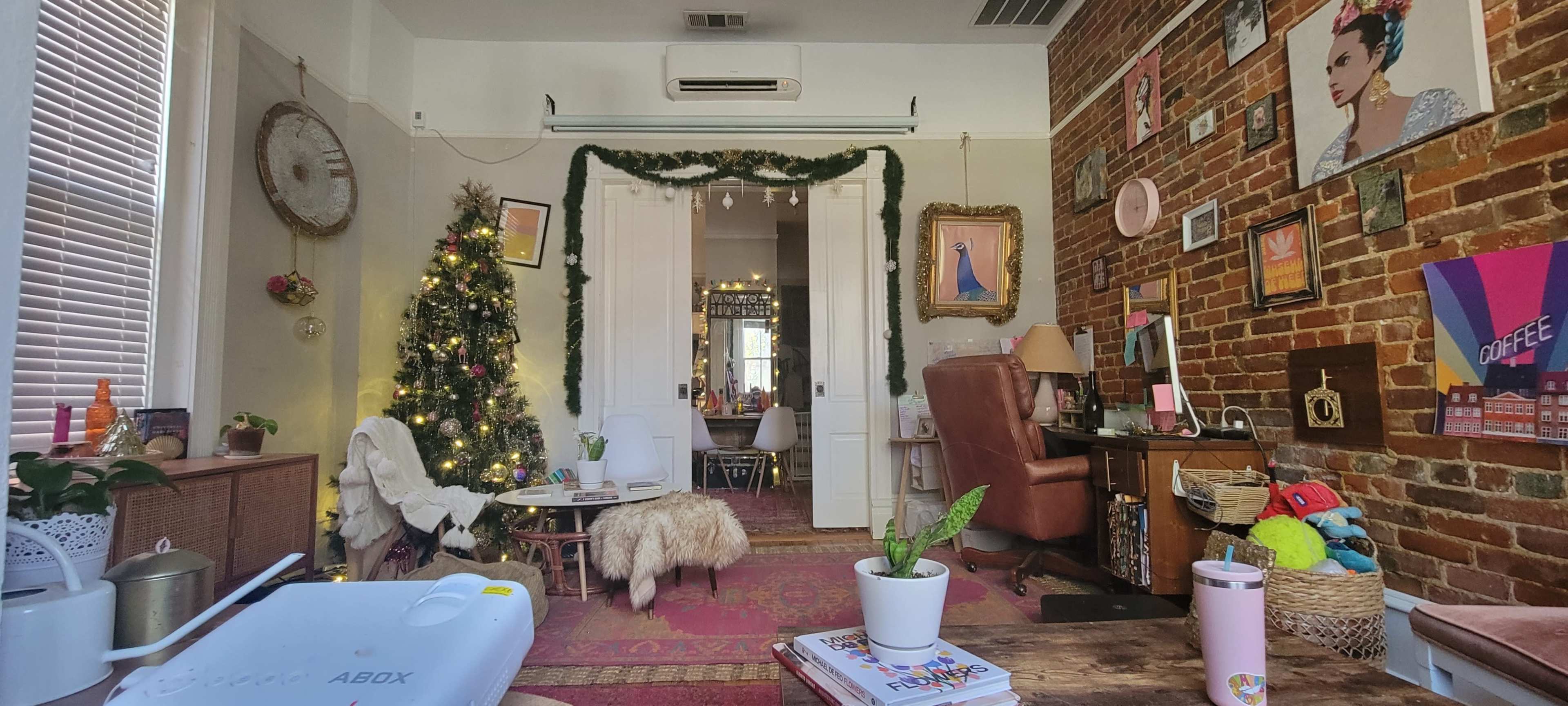 A cozy room with a decorated Christmas tree, a soft rug, a vintage armchair, and a workspace featuring a computer and various colorful decorations on the walls.