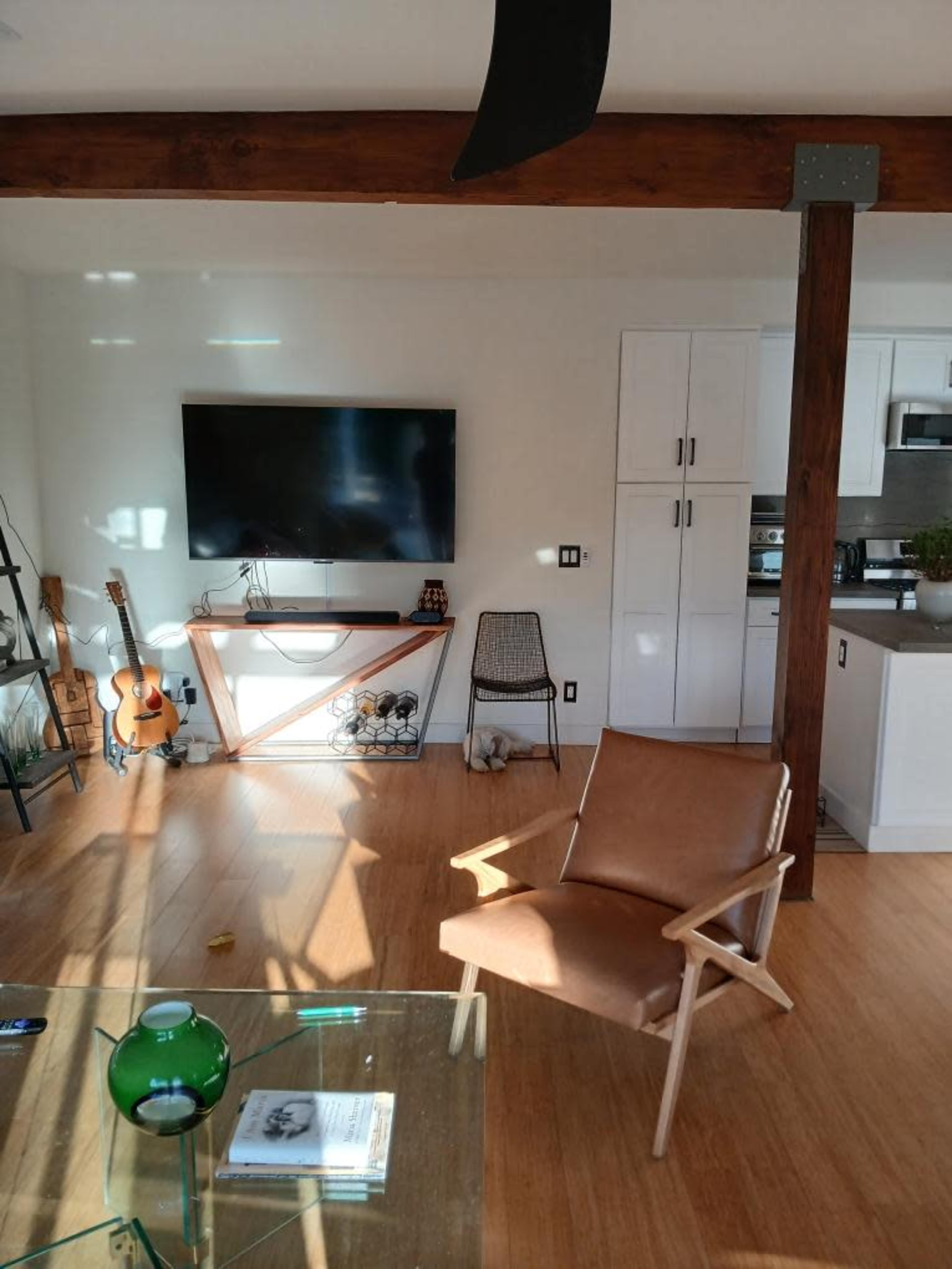 The image shows a modern living room with a wooden beam ceiling, a large TV mounted on the wall, a guitar, and a brown armchair positioned near a glass coffee table.
