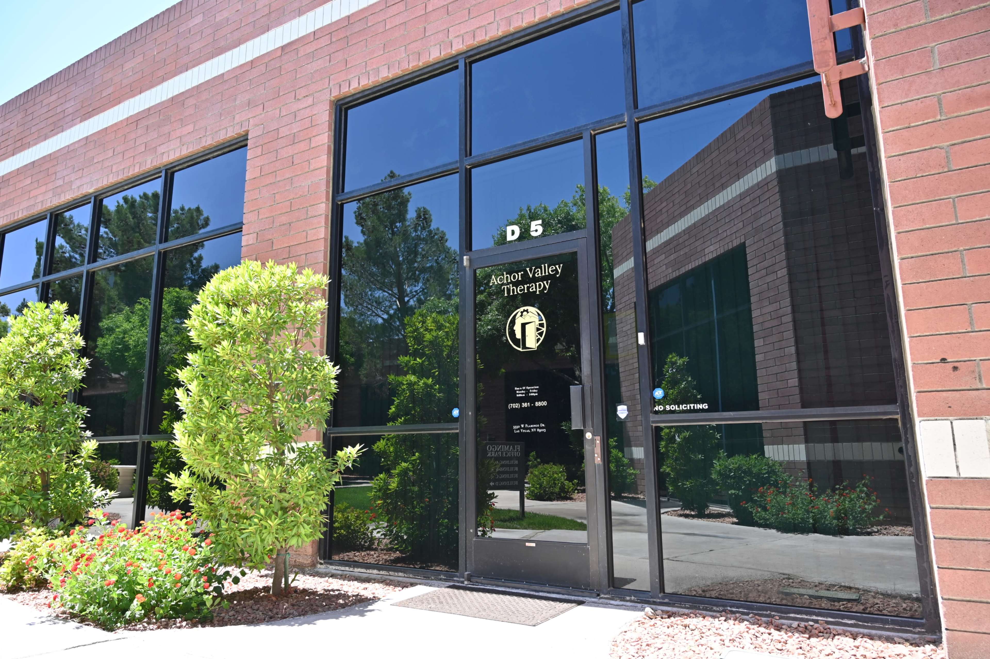 The image shows the entrance of Anchor Valley Therapy, located in a commercial building with large glass windows and surrounded by green shrubs and flowers.