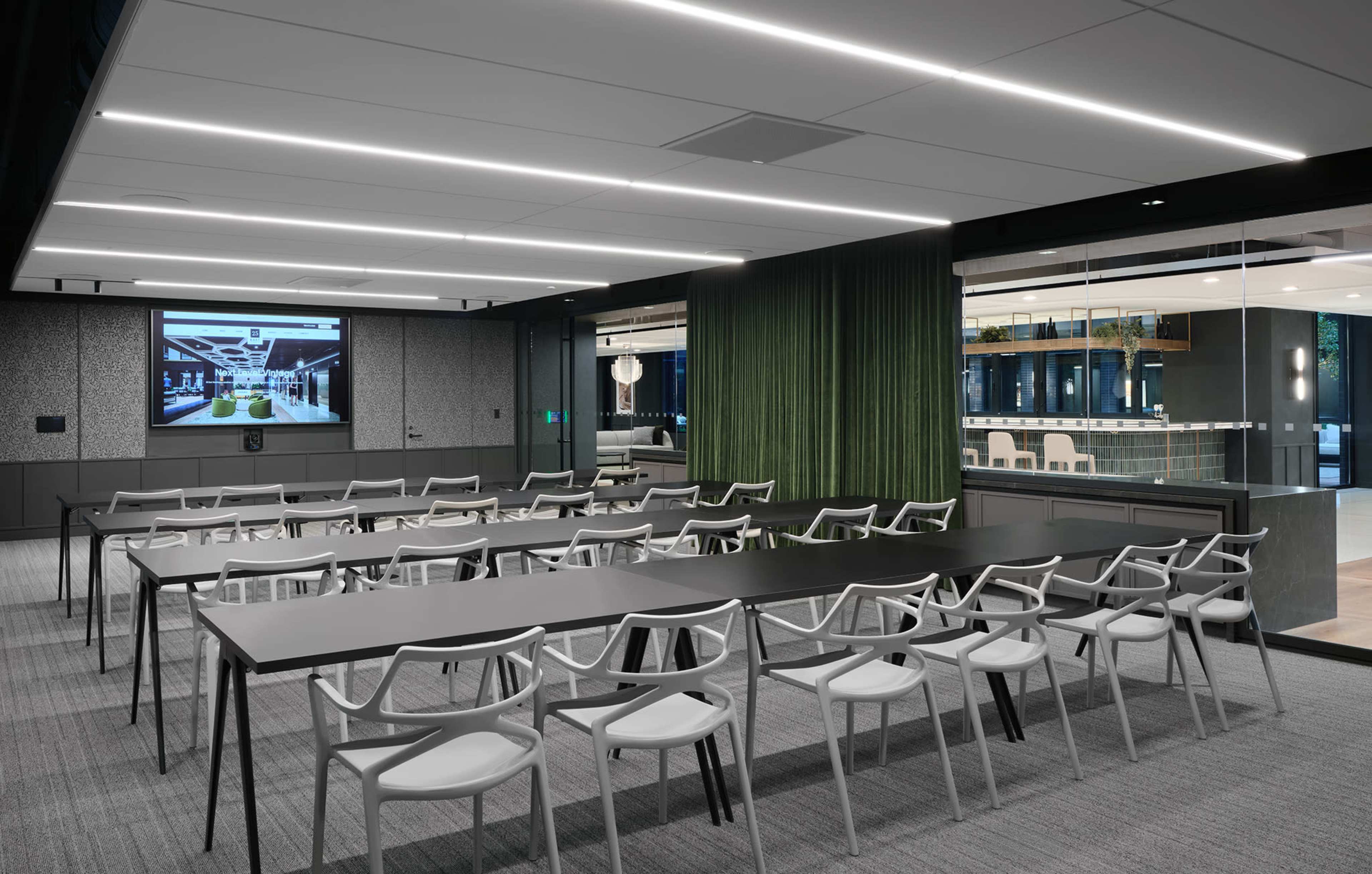 The image shows a modern conference room with a large screen at the front, lined with tables and chairs arranged for a meeting.