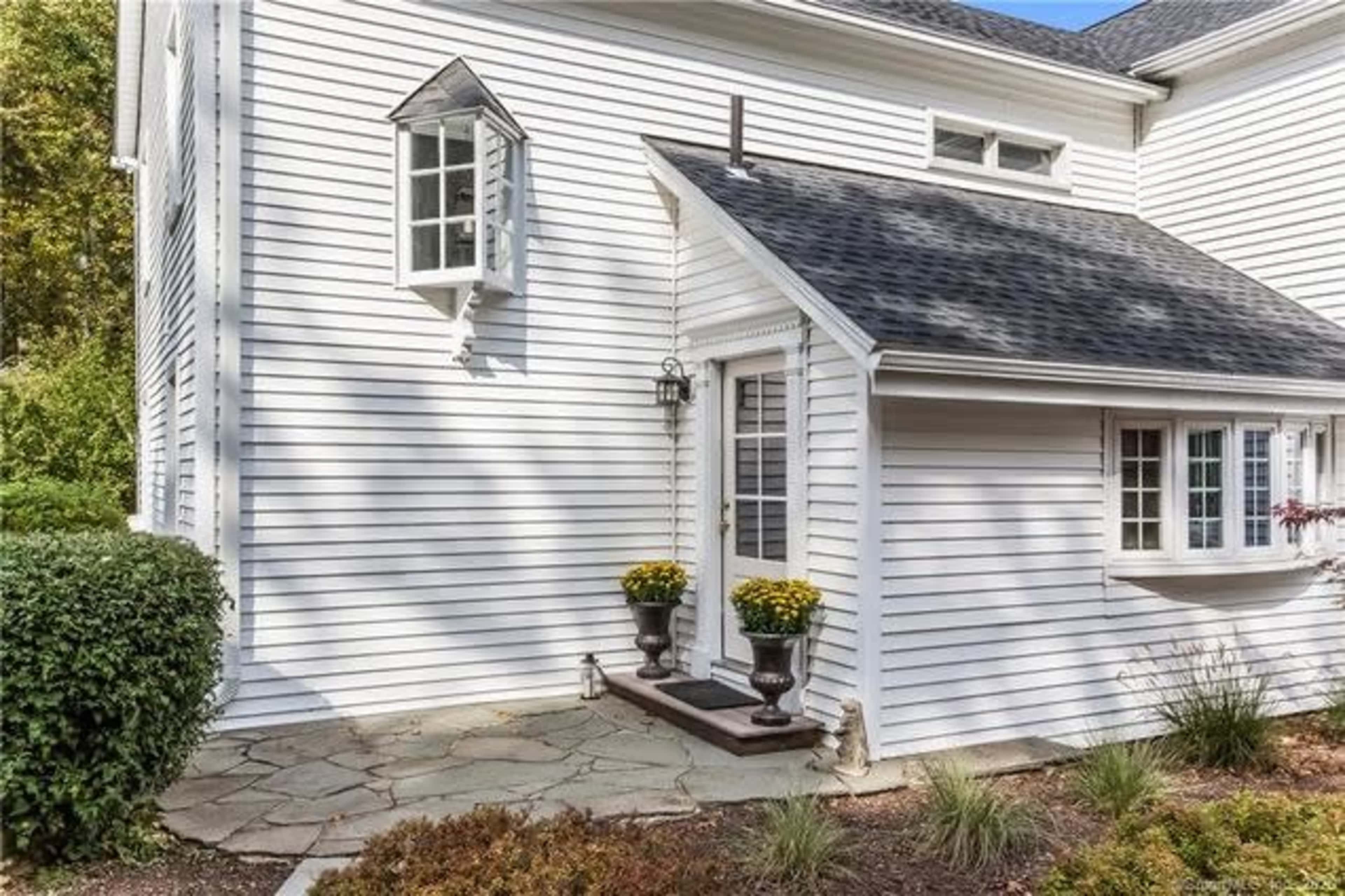 The image shows a corner of a house with white siding, featuring a small front porch with potted yellow flowers and a stone walkway.
