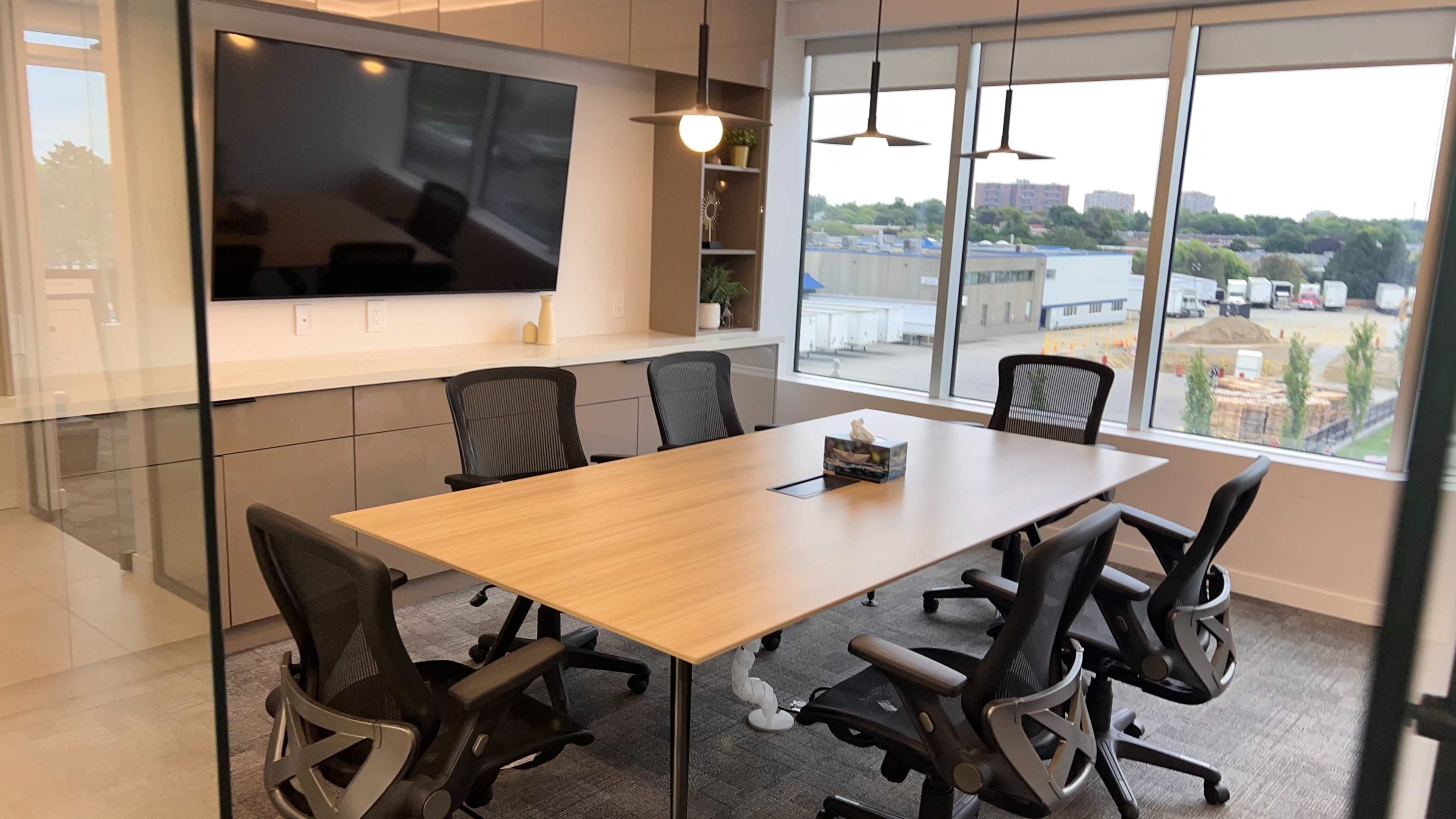 A meeting room features a large wooden table surrounded by six black office chairs, with a wall-mounted screen and large windows overlooking an outdoor area.