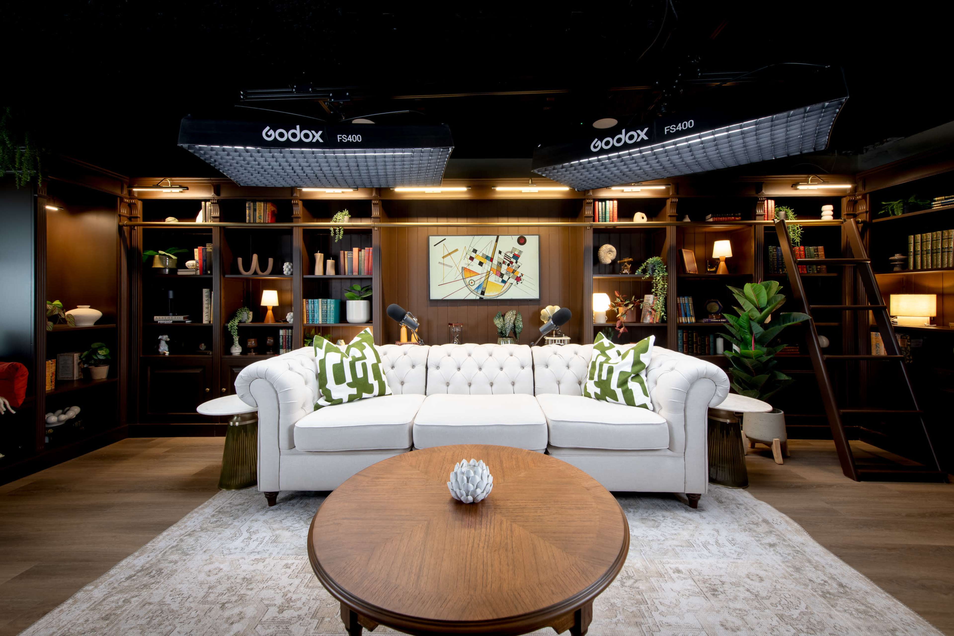 The image features a modern living room with a white tufted couch, a round wooden coffee table, and bookshelves filled with various items, illuminated by two overhead studio lights.