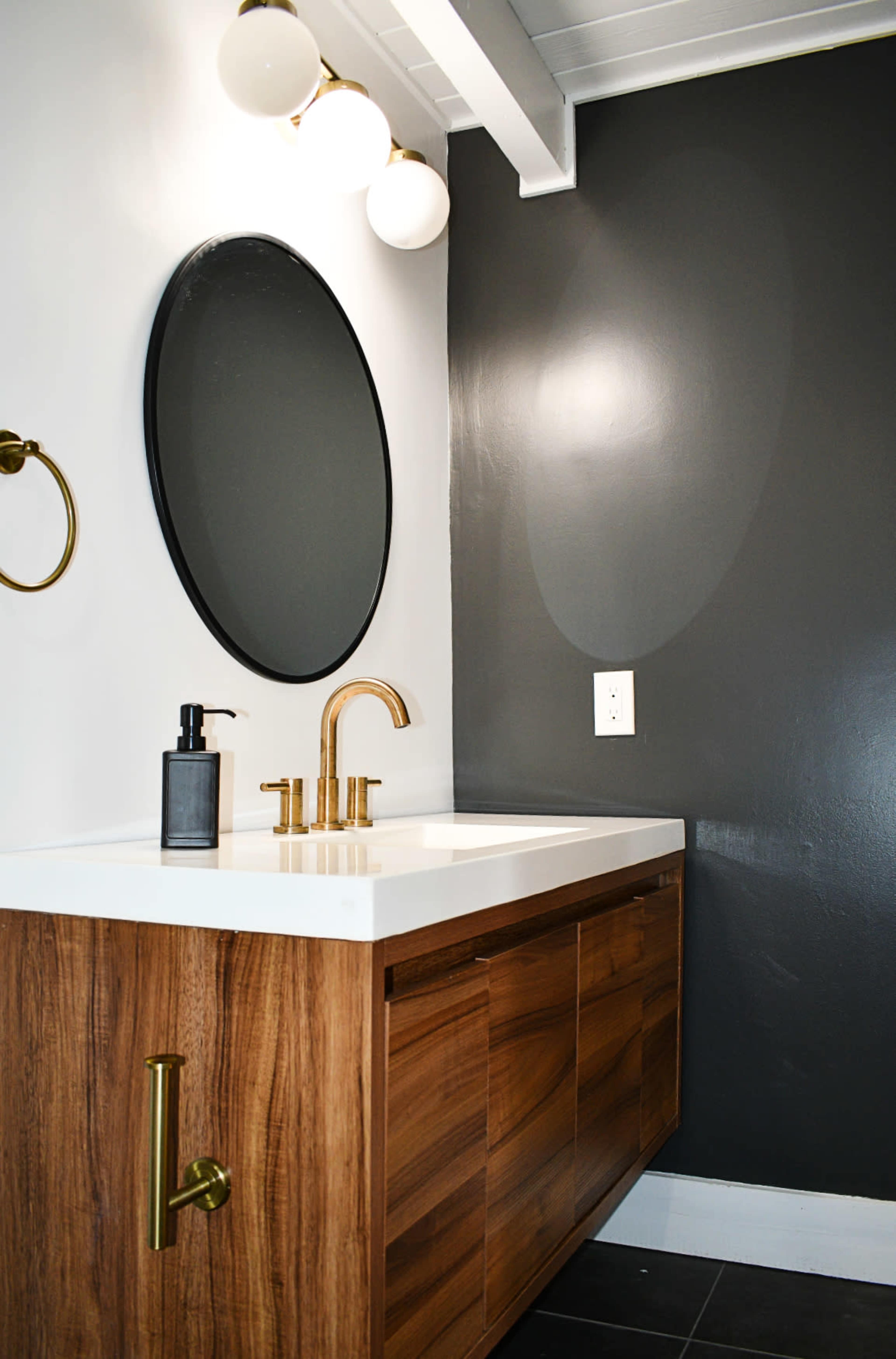 A modern bathroom with a circular mirror above a wooden vanity that features a white countertop, a gold faucet, and minimalist wall-mounted lighting.
