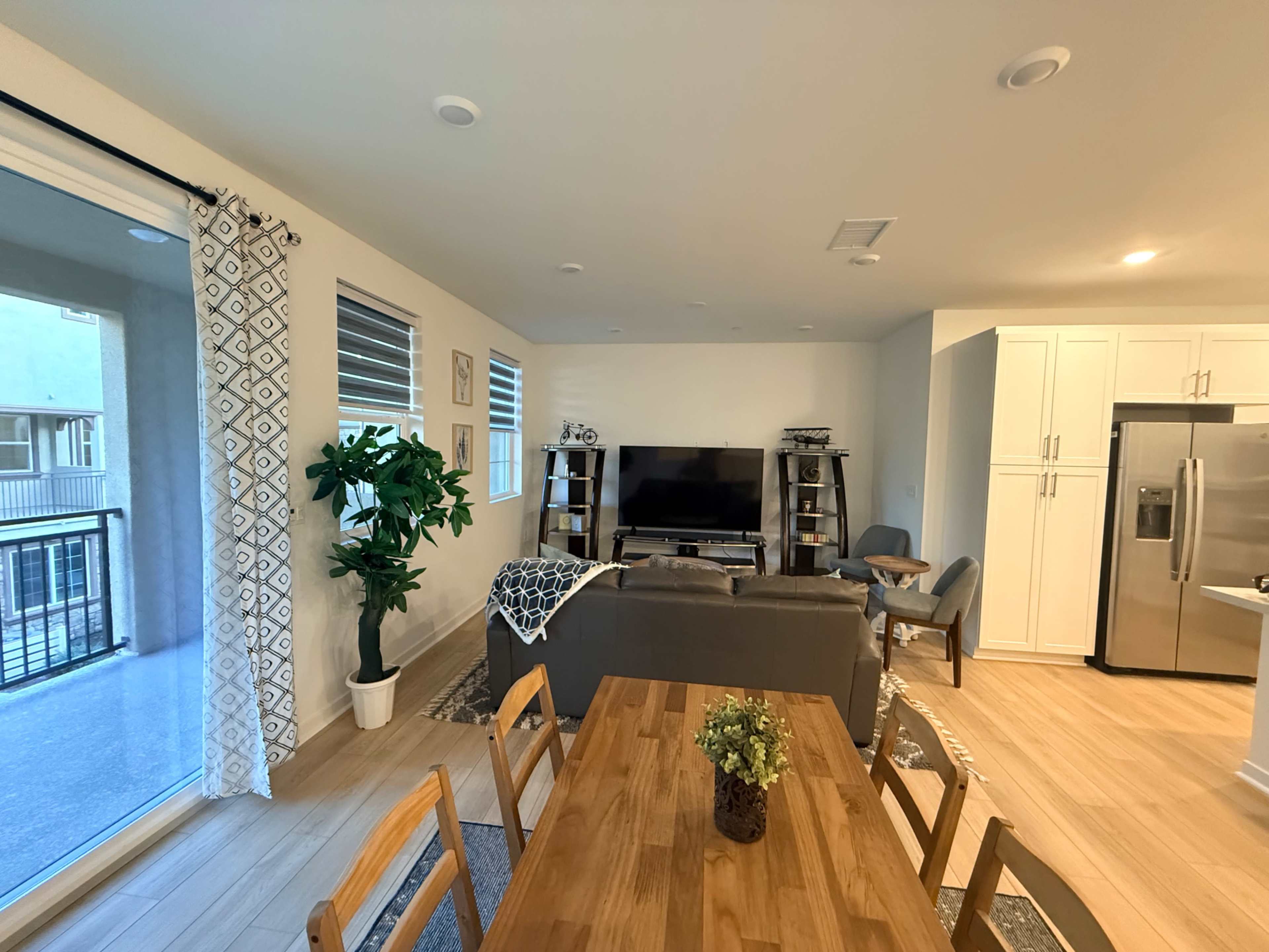 The image shows a modern living room and dining area with a couch, coffee table, TV, dining table, and large windows with sliding doors.