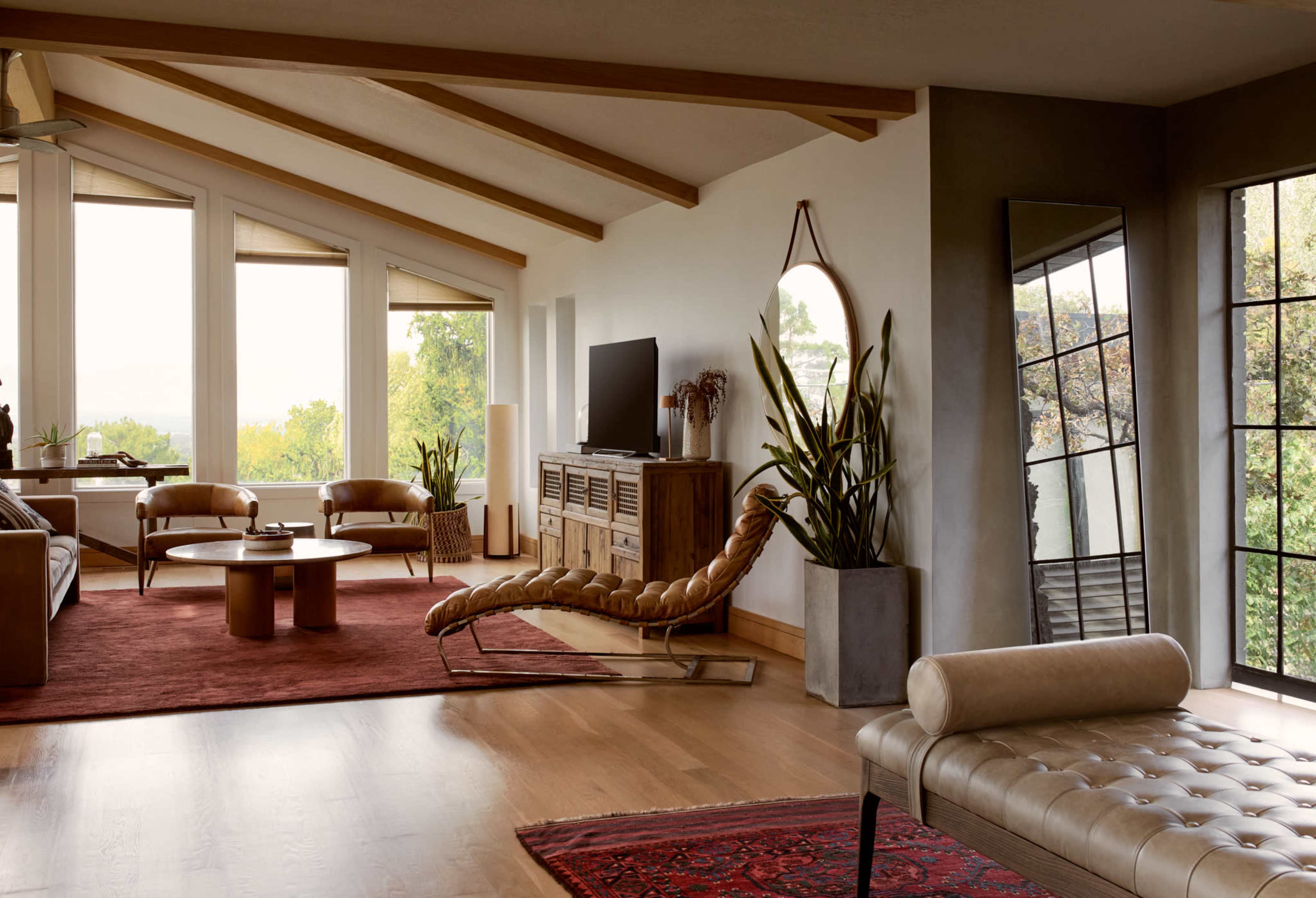 A modern living room features large windows, a wooden ceiling, a television console, and seating arrangements with a rug and plants.