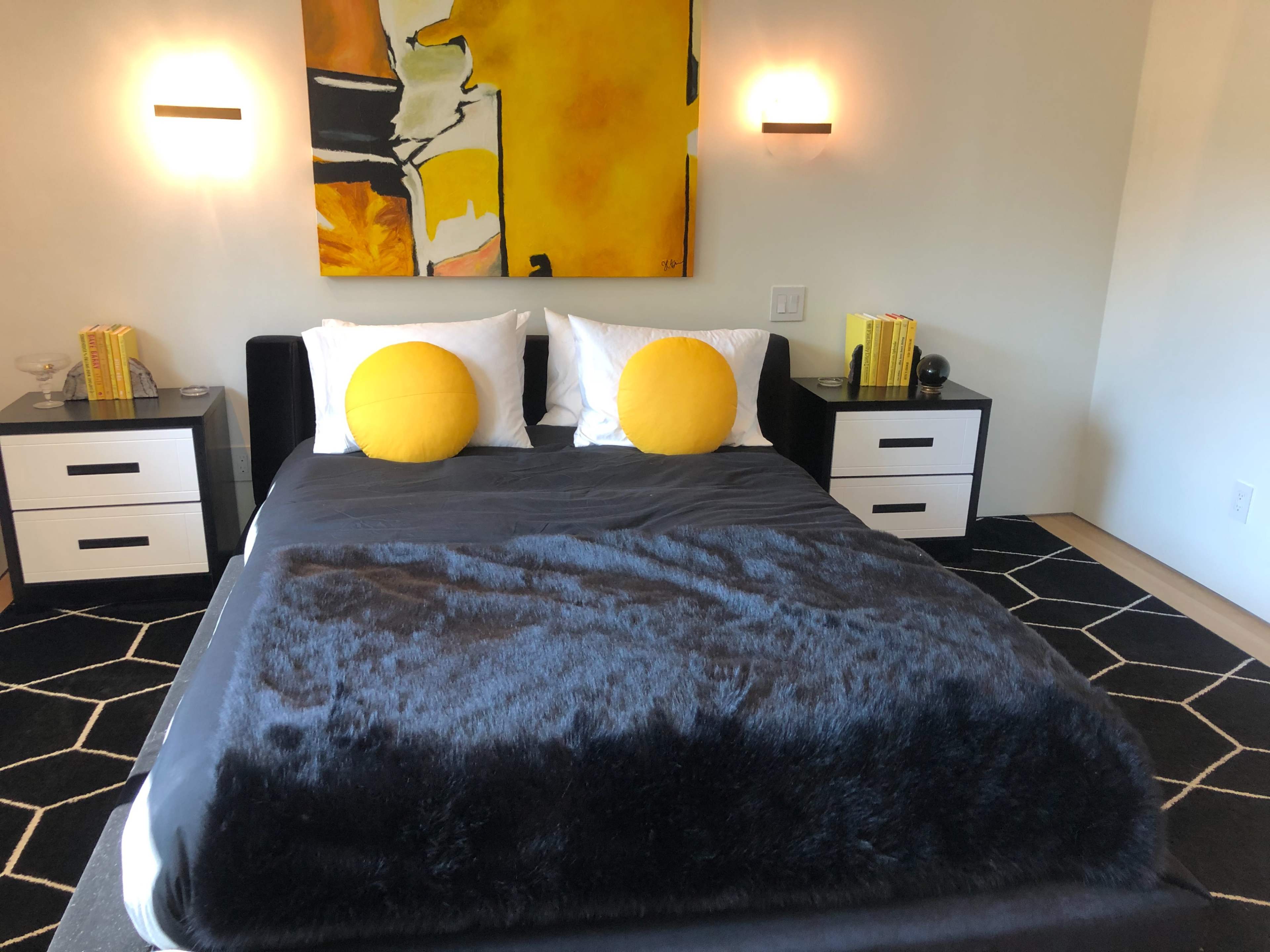 A well-lit bedroom features a black bedspread, two yellow accent pillows, and nightstands with decorative items on either side.