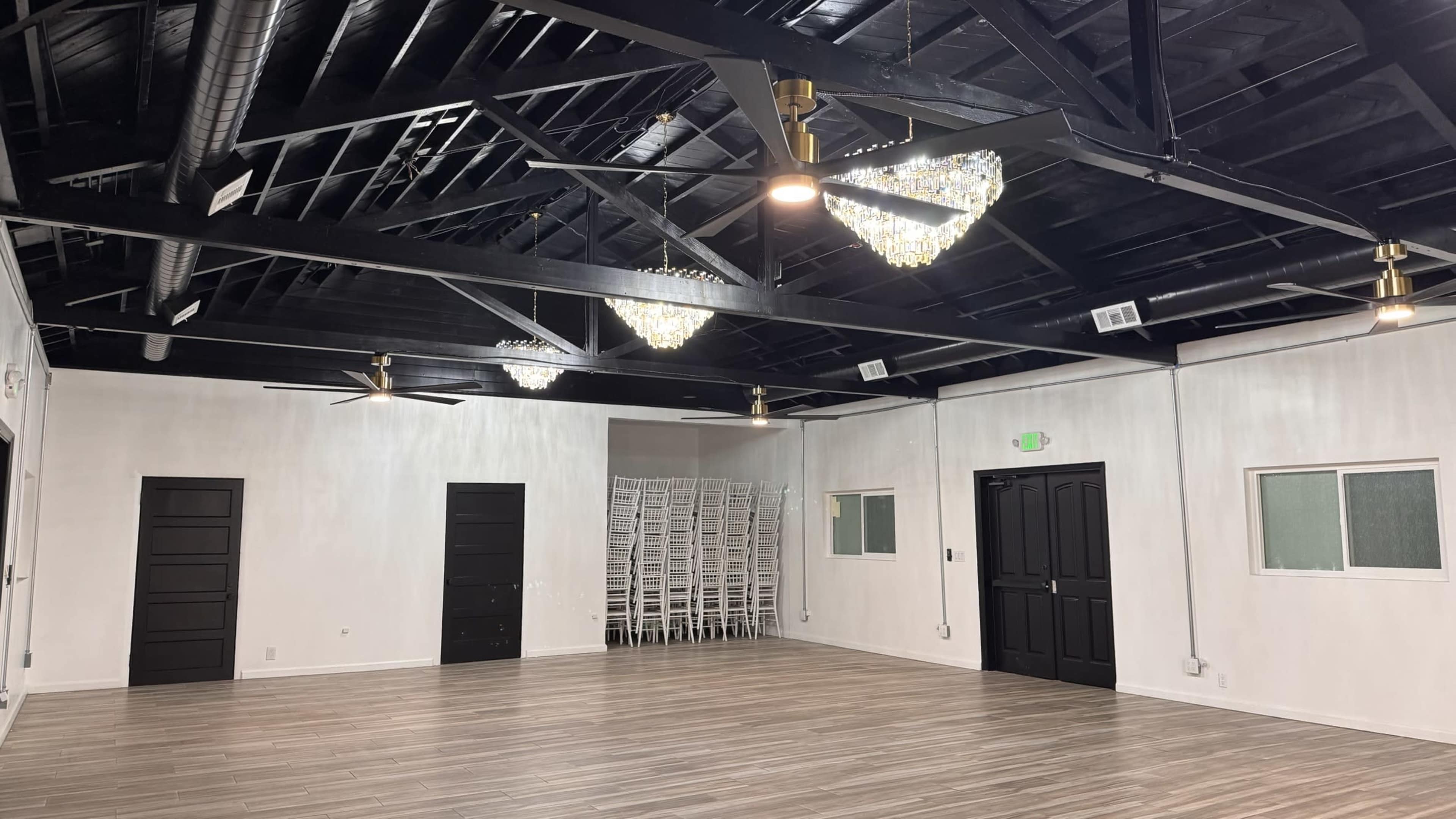 The image shows an empty event space with a polished wood floor, black ceiling beams, and three decorative light fixtures.