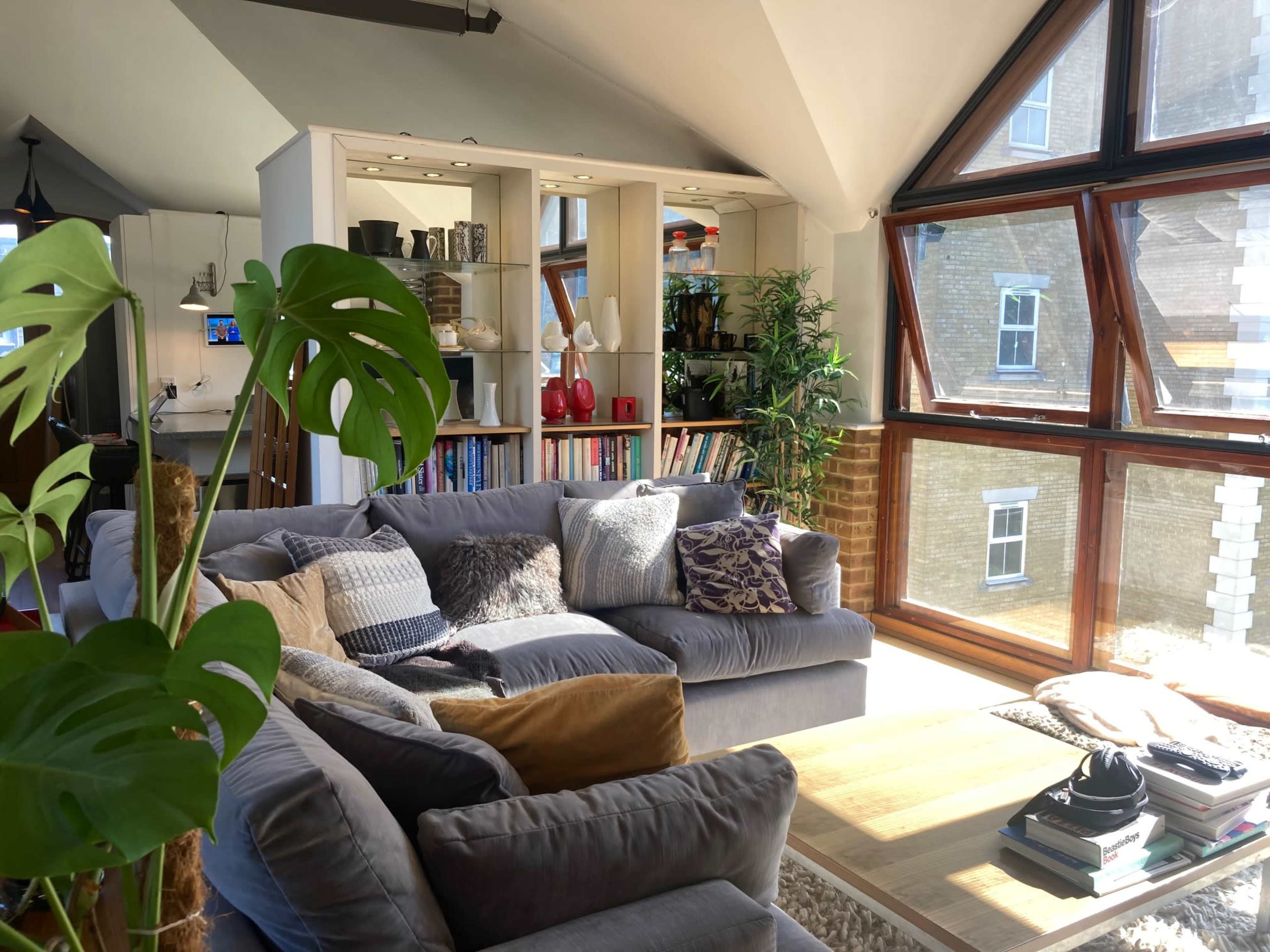 A spacious living room features a large sectional sofa, a wooden coffee table, and a bookshelf filled with colorful decor, all illuminated by natural light from tall windows.