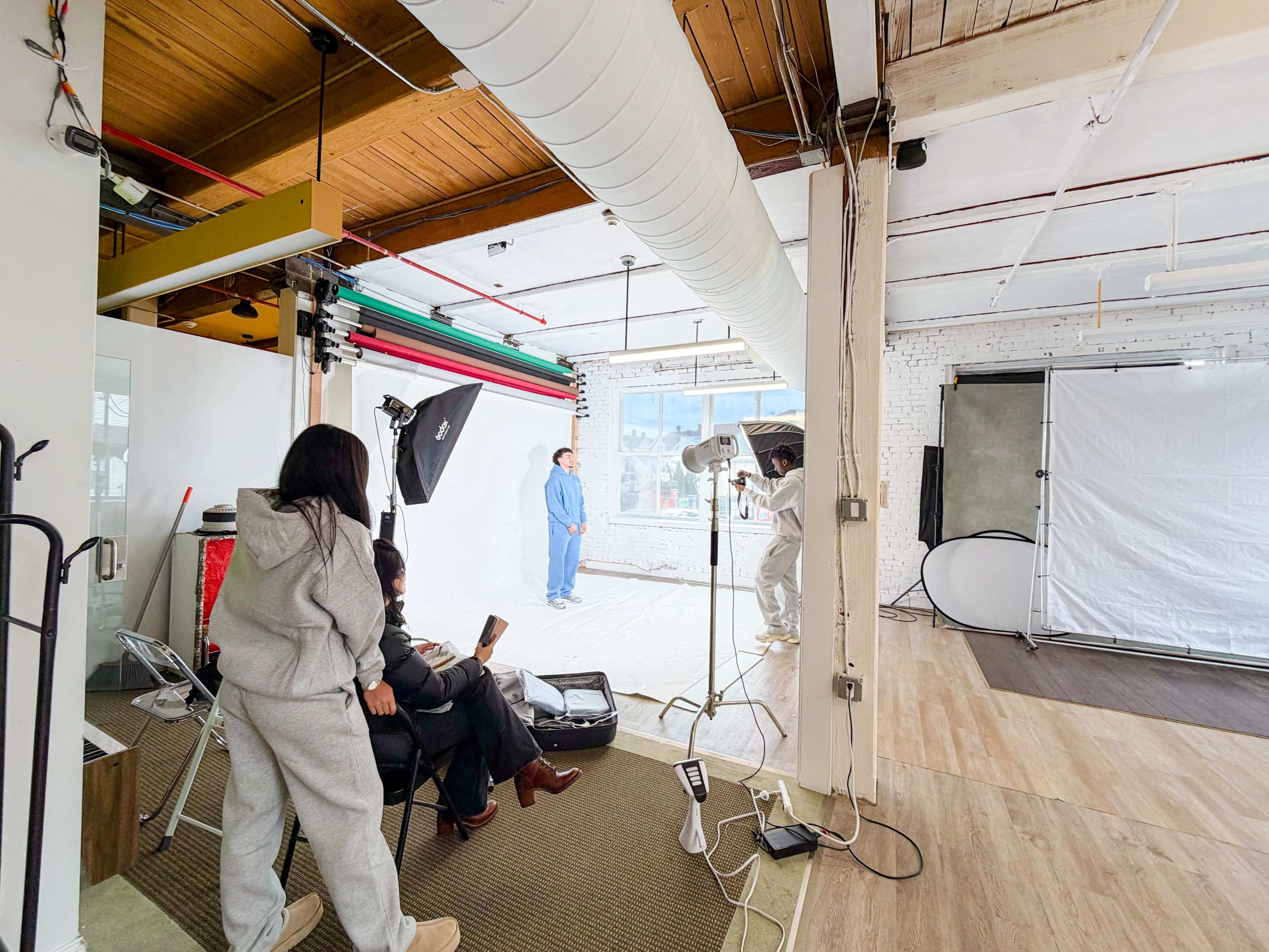 A photography studio features a subject posing in the background while a few individuals observe and prepare equipment in the foreground.