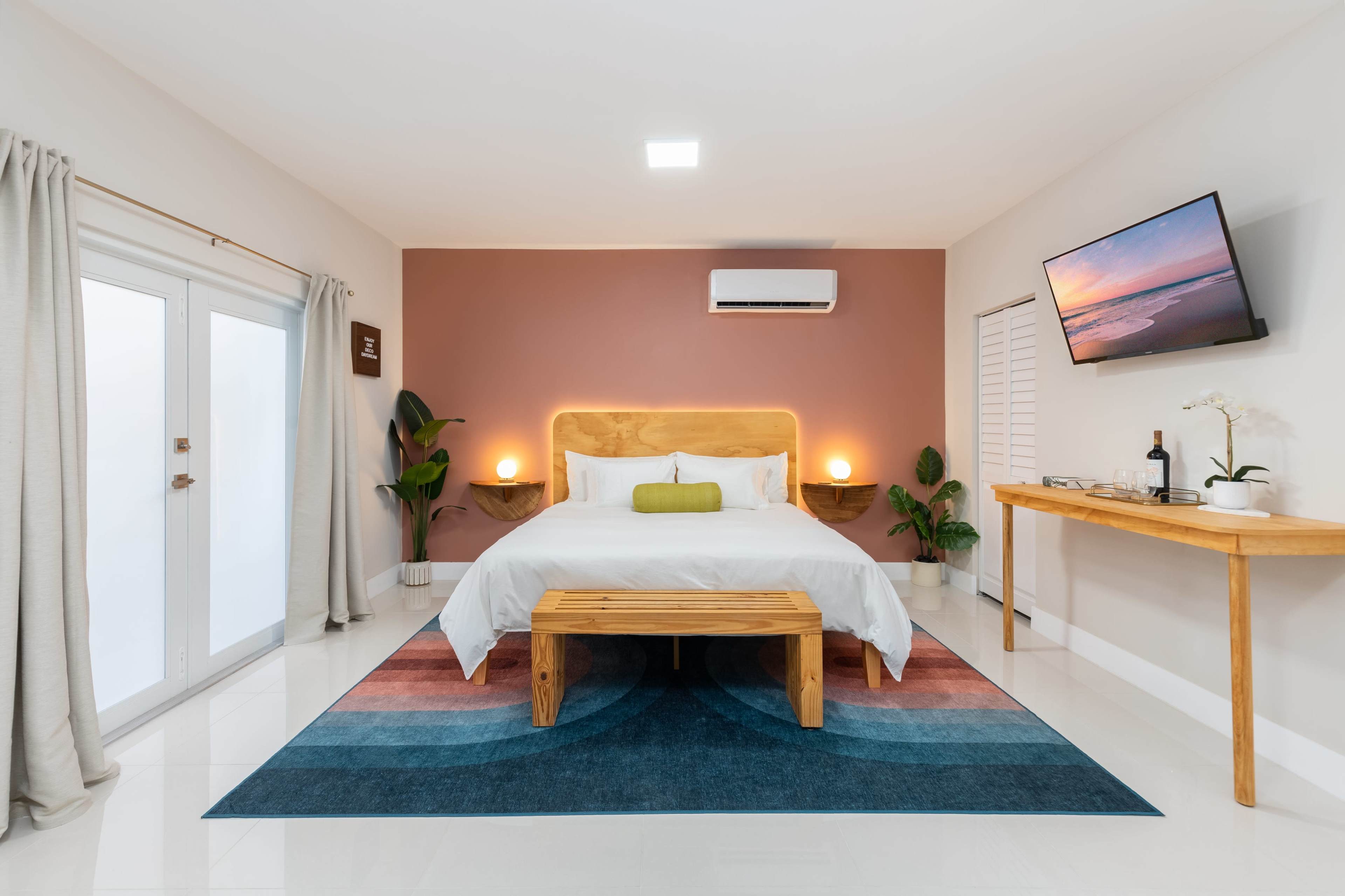 A modern bedroom features a wooden bed with a green pillow, a bench at the foot, a colorful rug, and a wall-mounted TV.