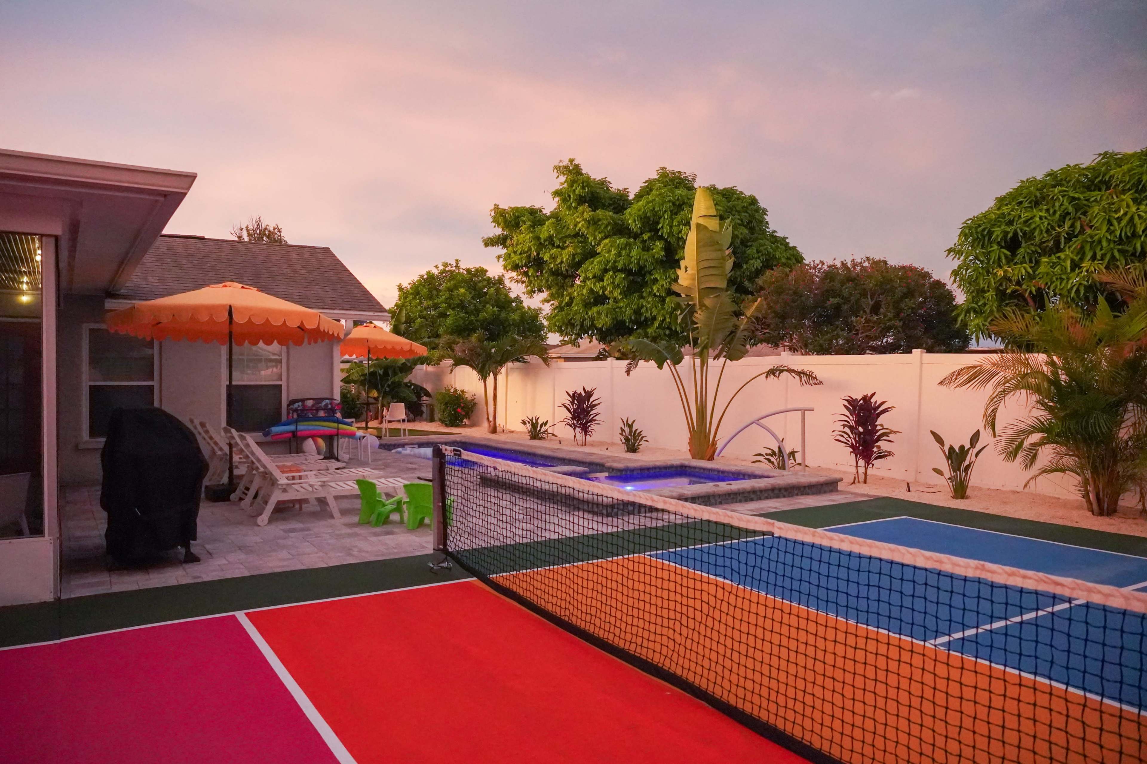 The scene features a backyard with a tennis court, swimming pool, and landscaped greenery, illuminated by a sunset sky.