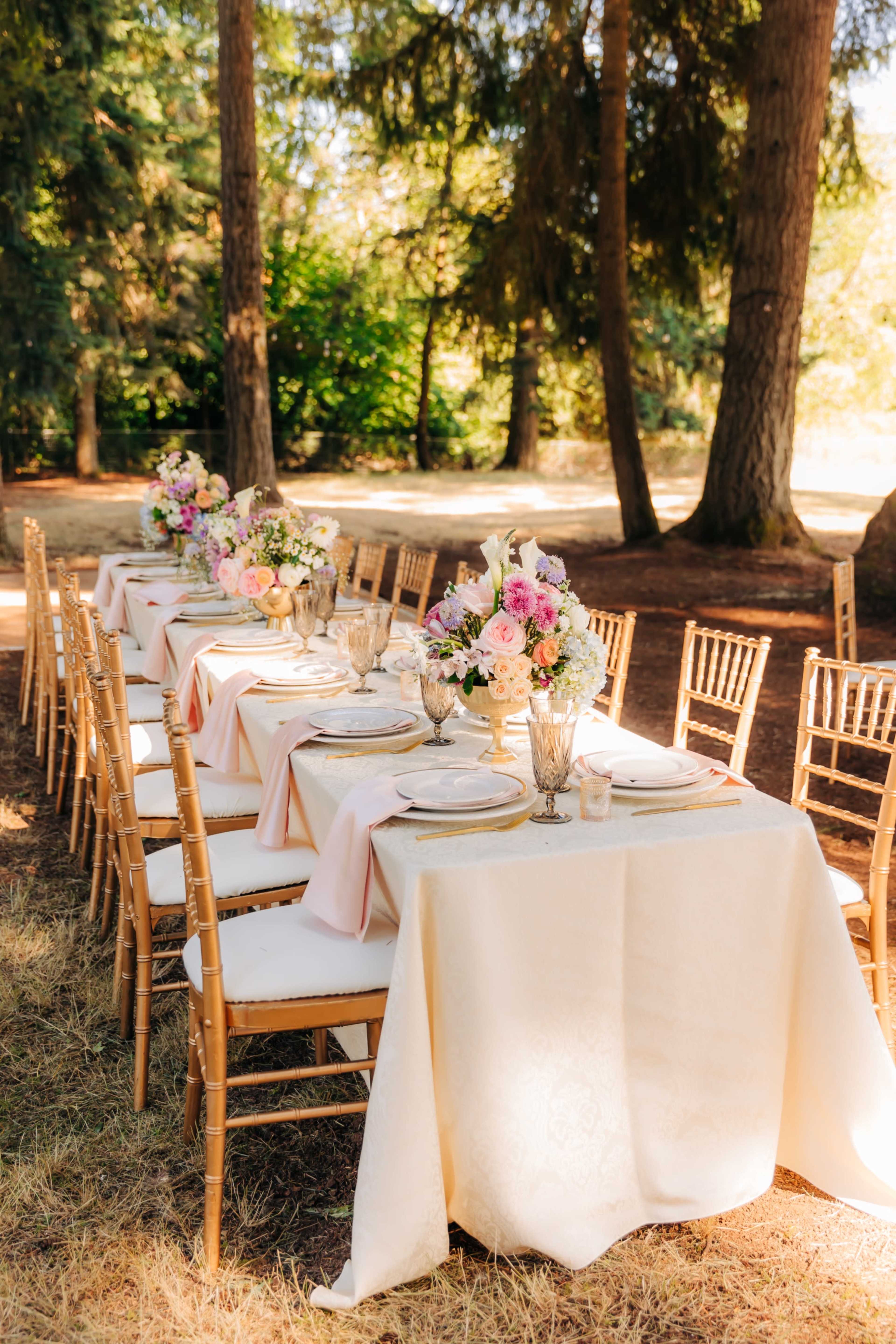 A long banquet table is set outdoors, adorned with floral centerpieces, plates, and glassware, surrounded by wooden chairs under tall trees.