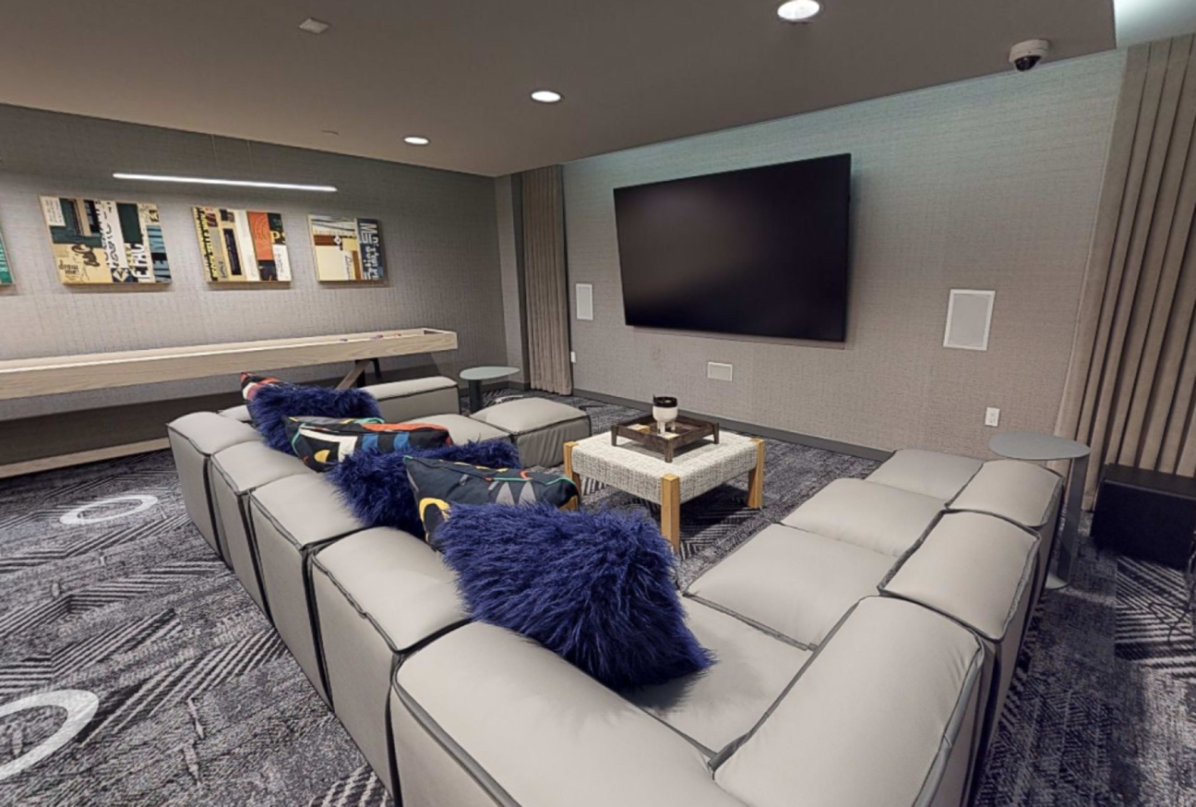 The image shows a modern lounge area with a large flat-screen TV mounted on the wall, surrounded by a gray sectional sofa and three small side tables.