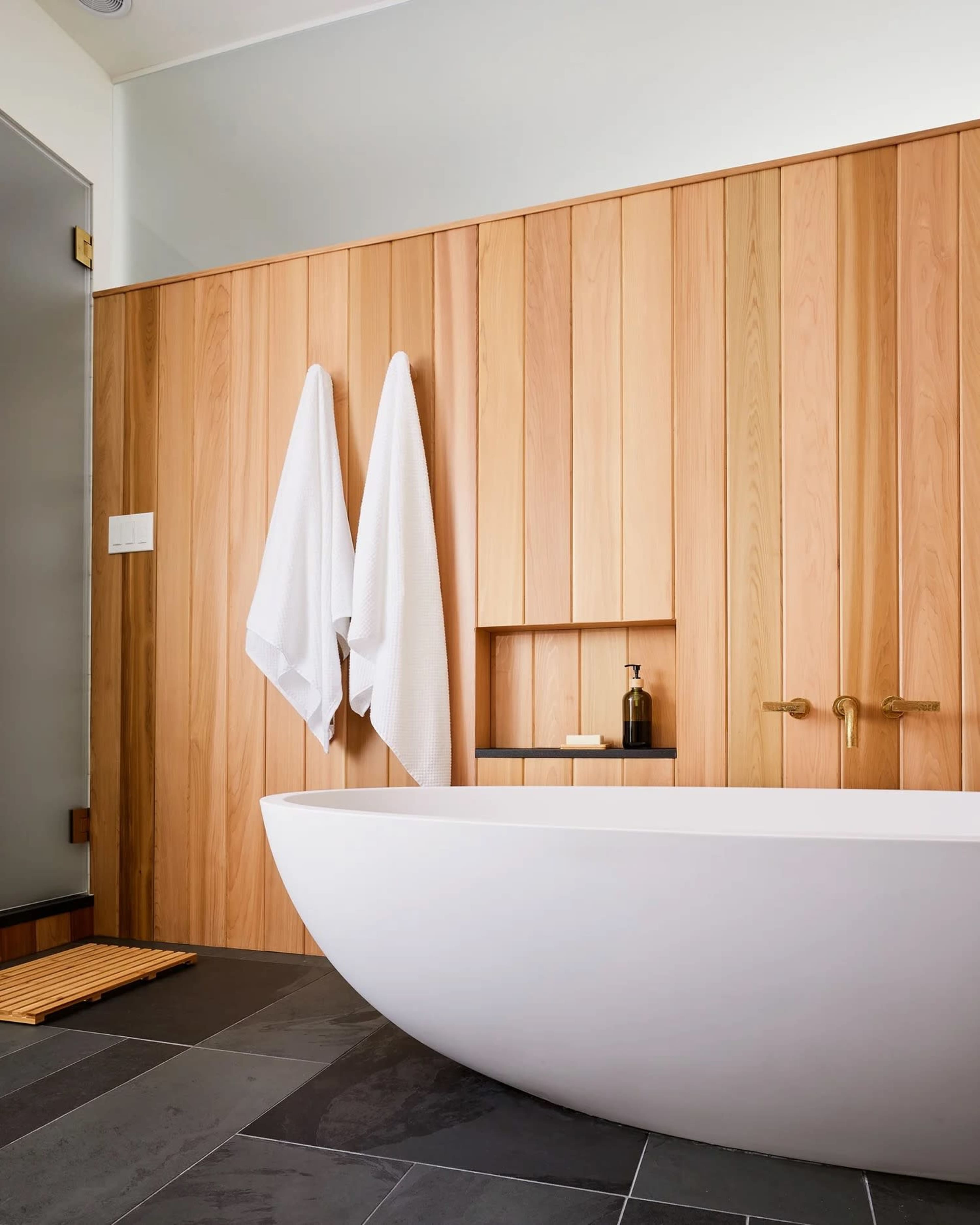 A modern bathroom features a freestanding white bathtub against a wooden accent wall with hanging towels and a shelf.