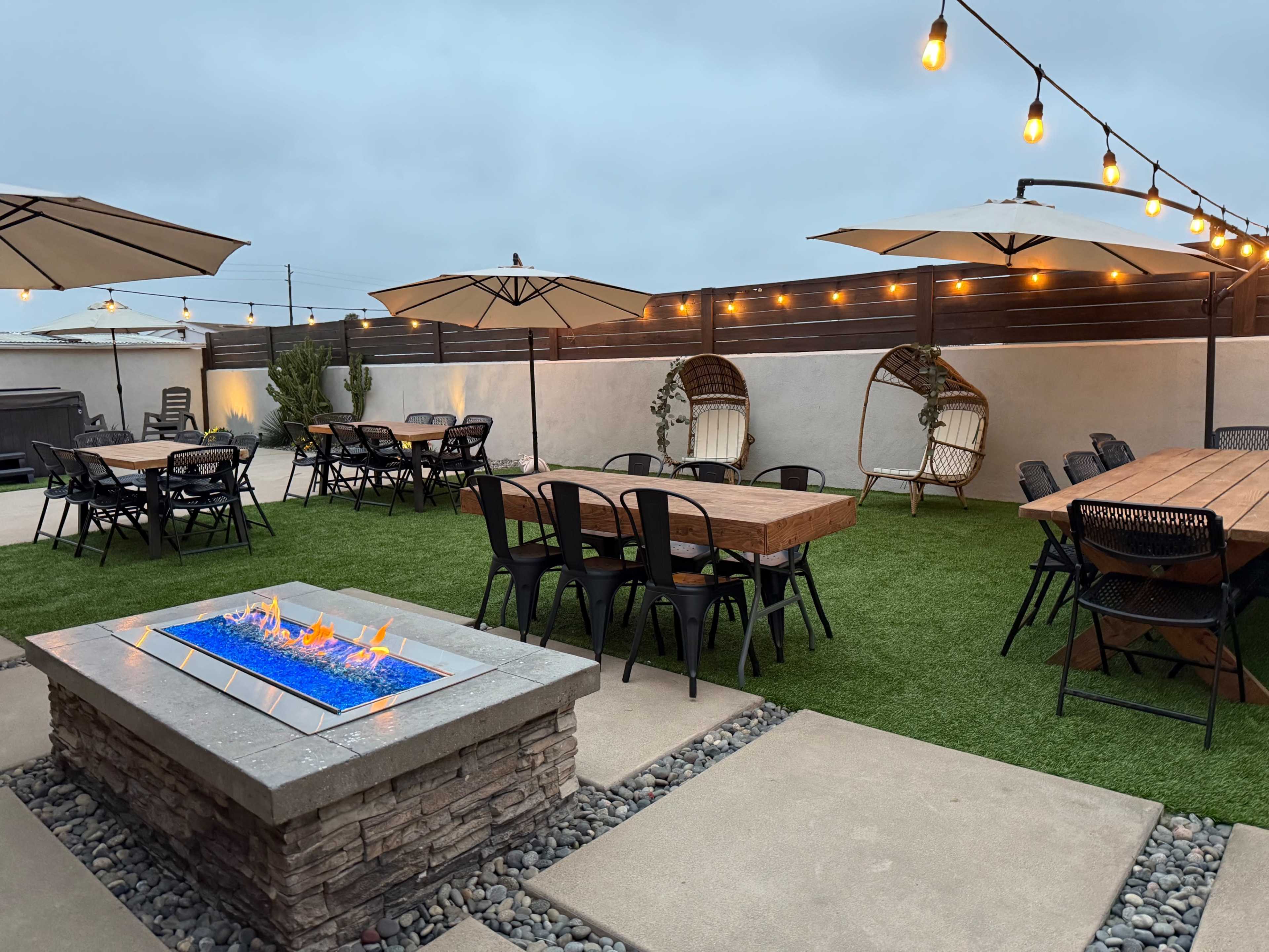 The image shows an outdoor patio area featuring several dining tables, umbrella shades, a fire pit with flames, and string lights overhead.