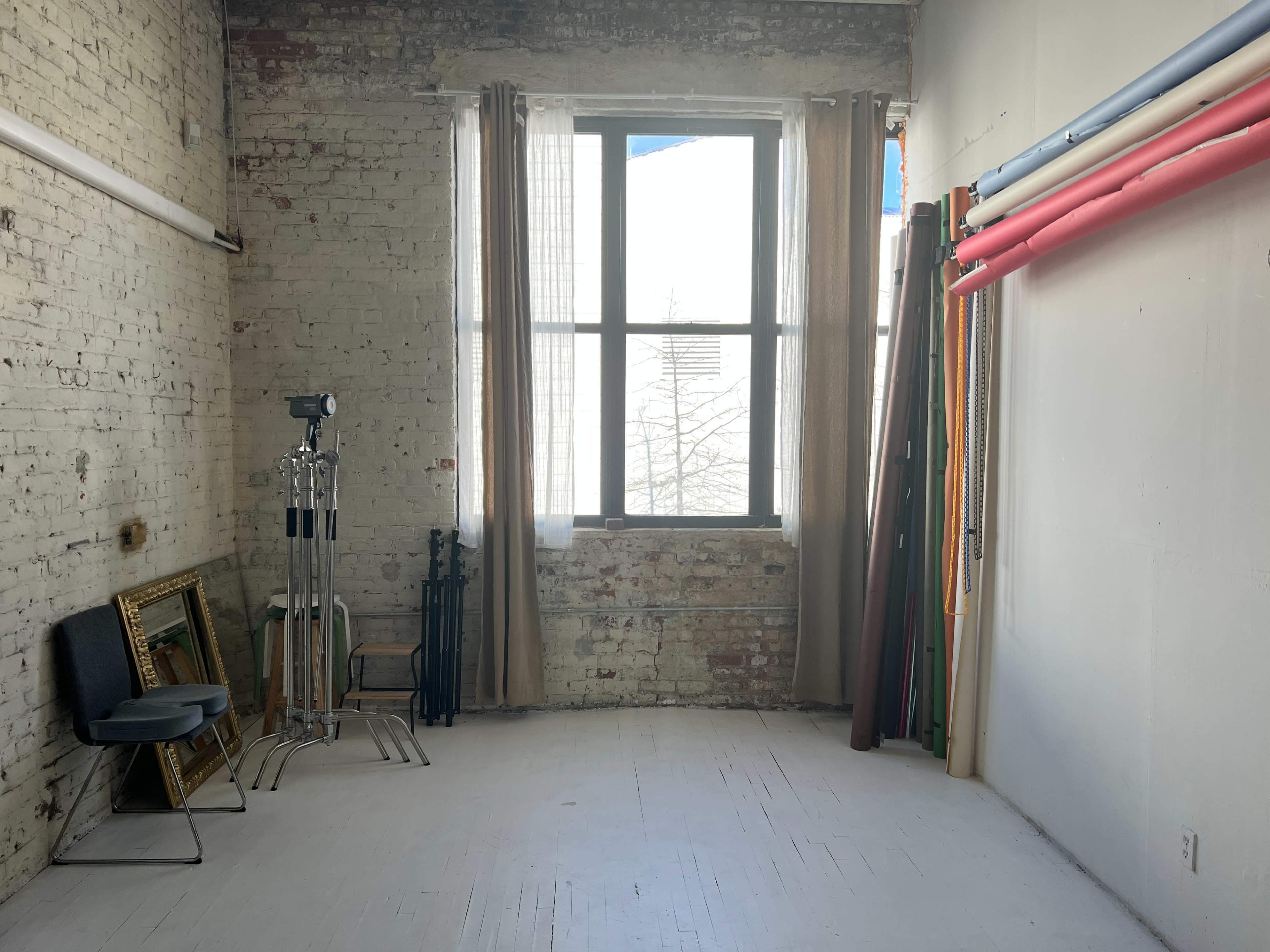 A spacious room with exposed brick walls, a tall window, and various props including a chair and colorful backdrops.