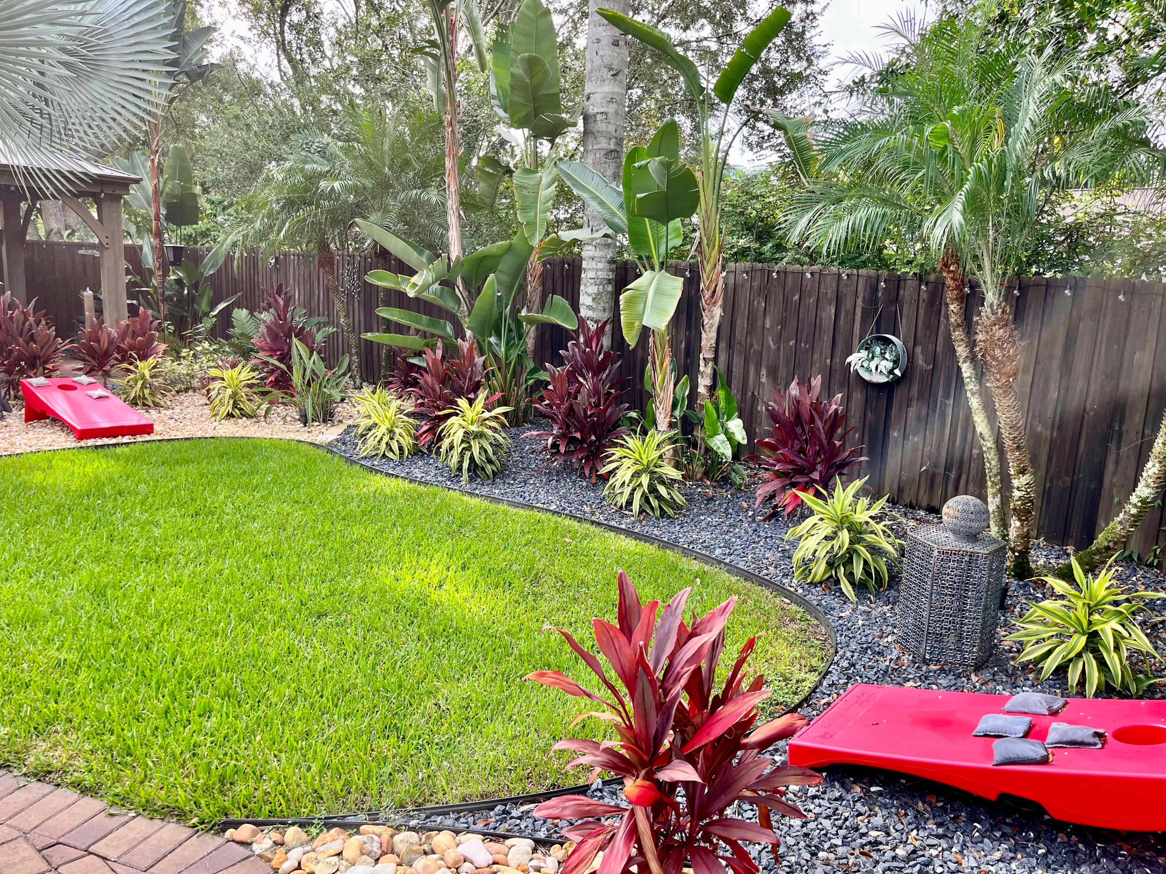A landscaped backyard featuring a green lawn, decorative plants along the edges, and two red cornhole boards set up on the grass.