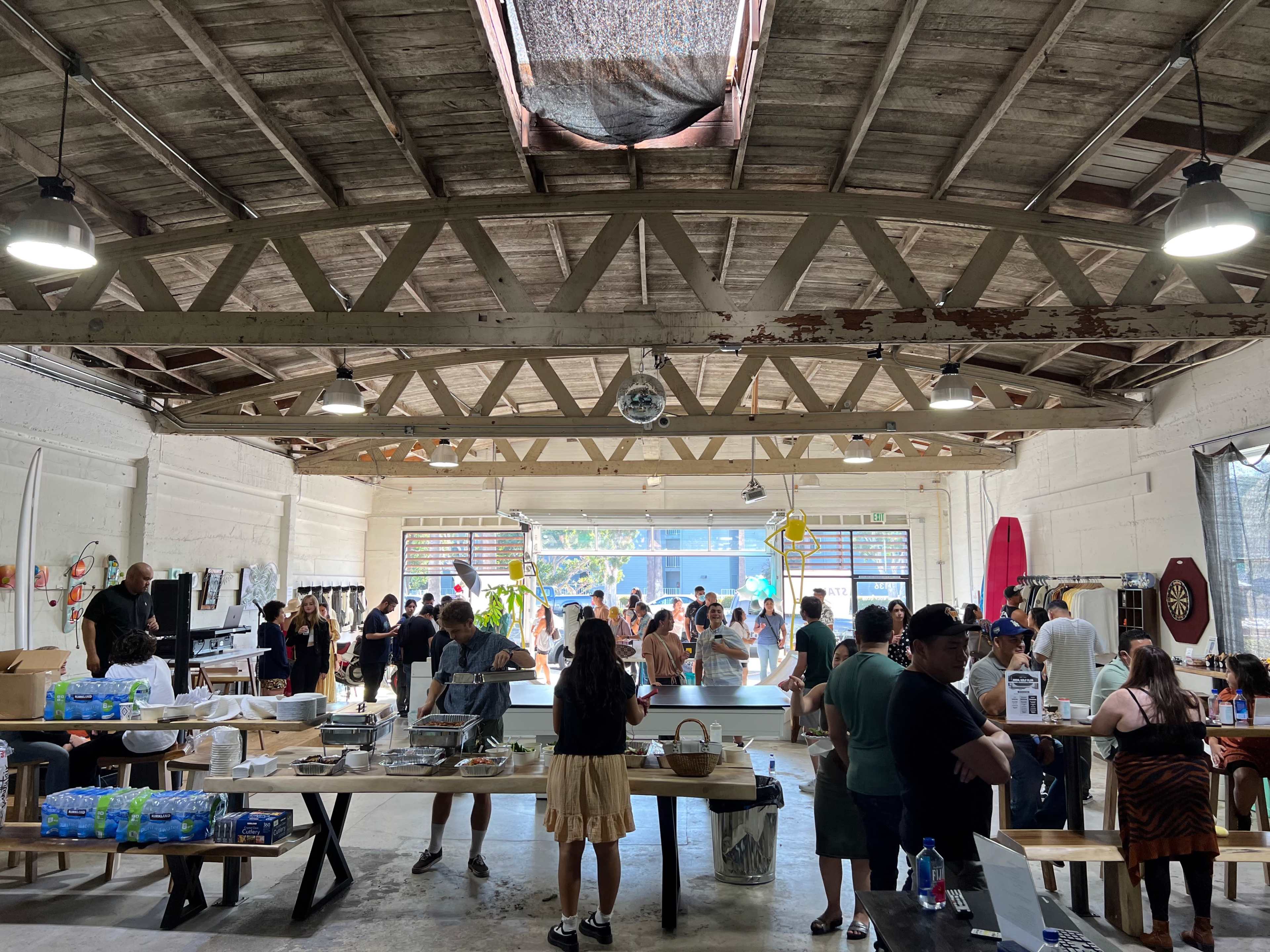 A large, open space filled with people engaging in various activities, including tables with food and drinks, surfboards on display, and natural light streaming through windows.