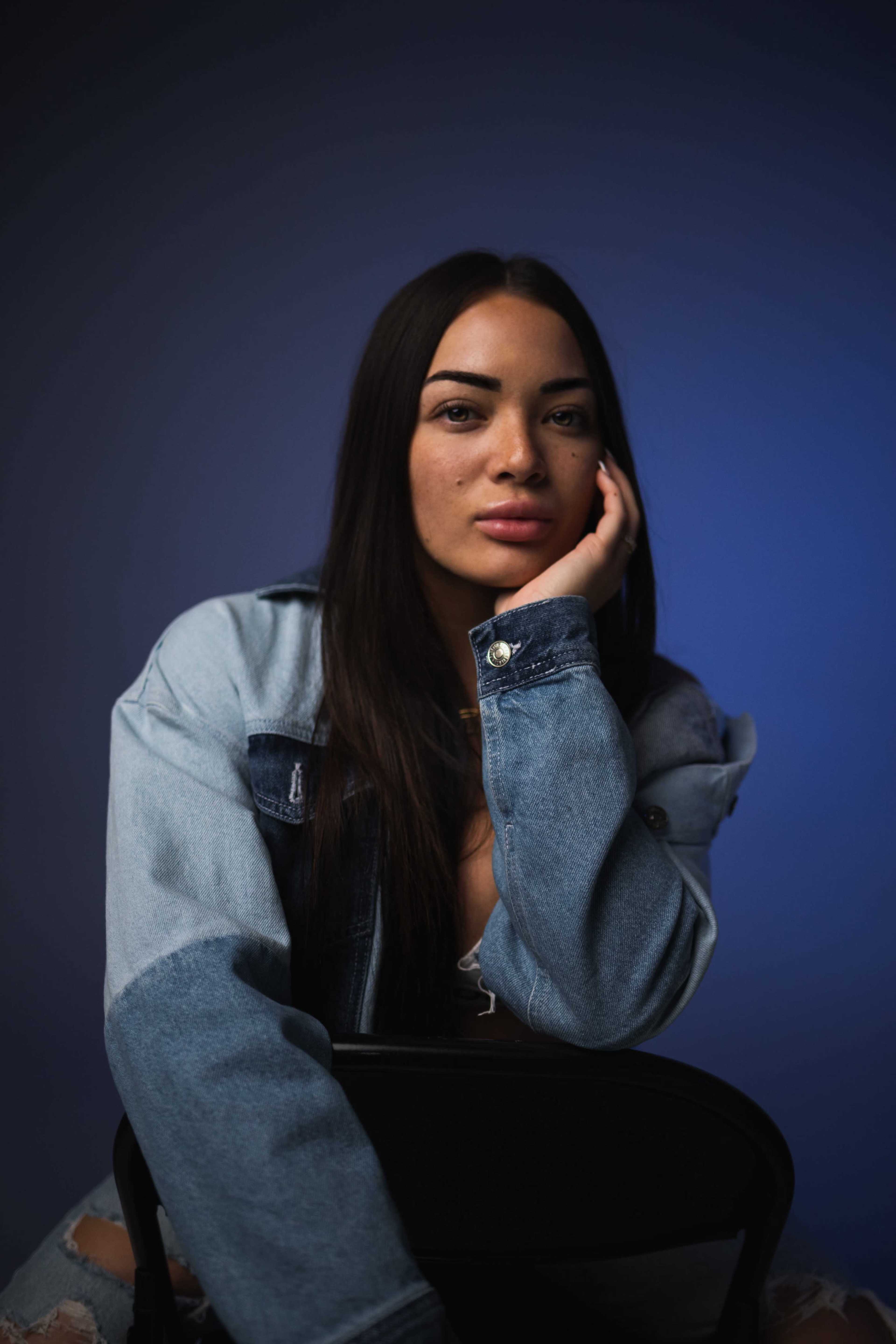 A woman with long hair rests her chin on her hand while seated against a blue background.