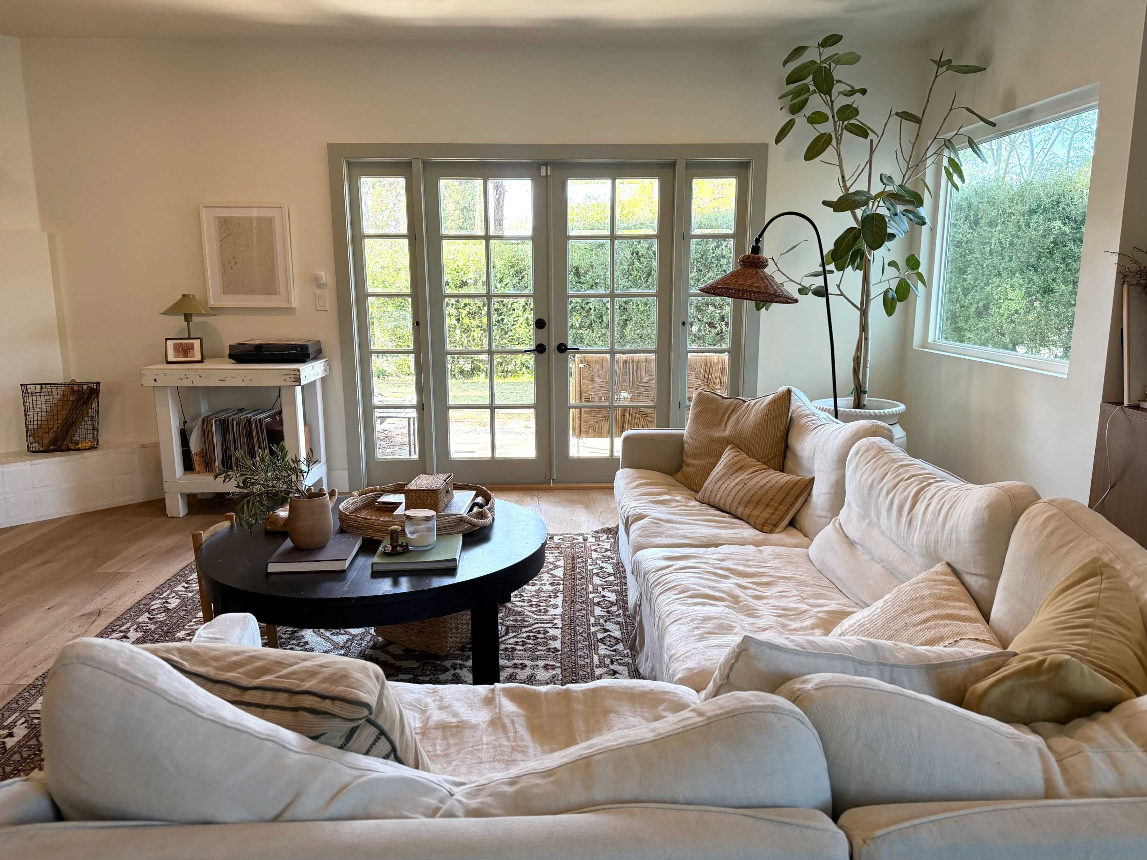 A light and airy living room features a large white sectional sofa, a round black coffee table, and glass doors opening to a backyard garden.