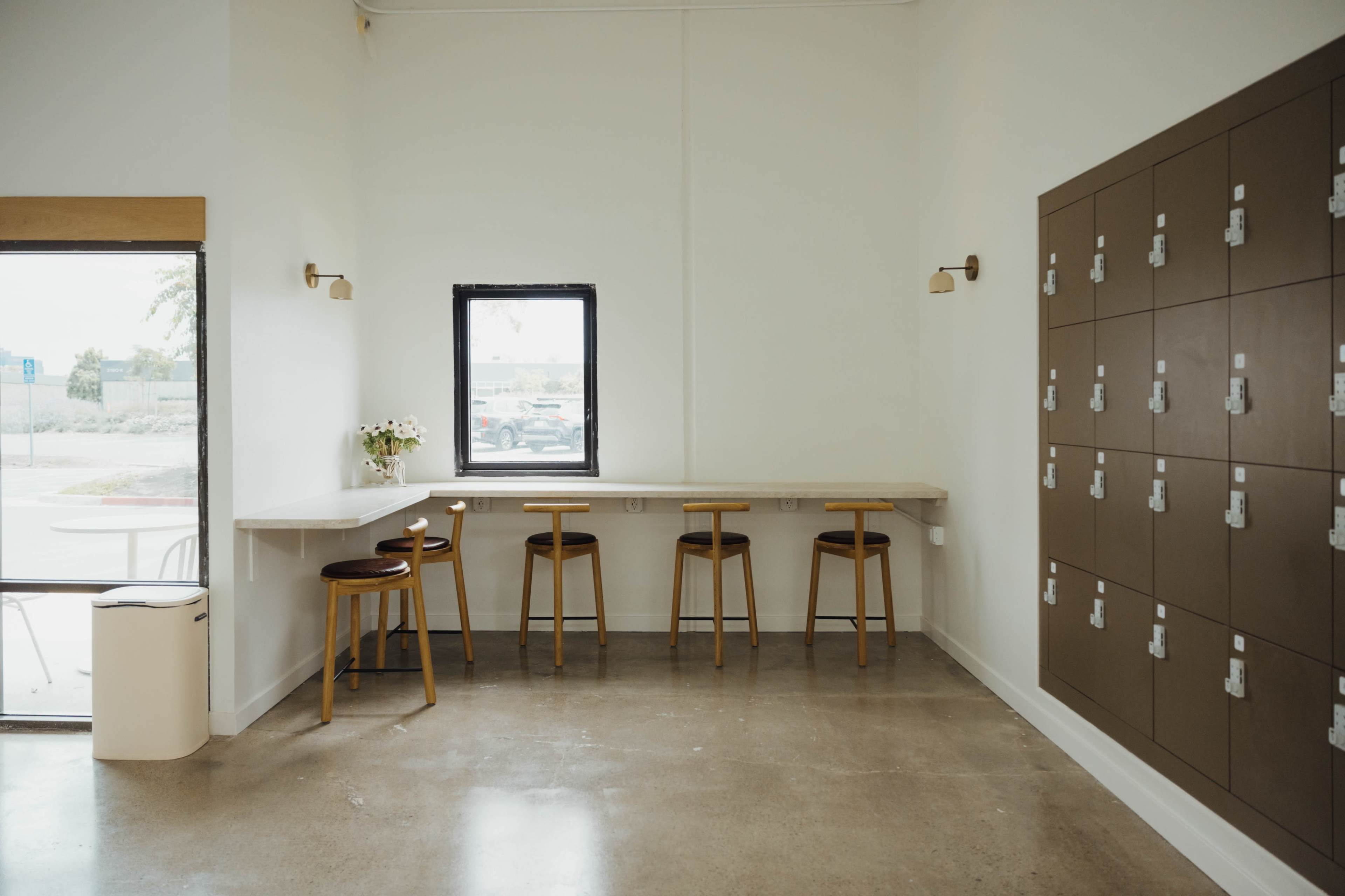 The scene features a minimalist interior with a long counter and four wooden stools beside a wall of lockers.