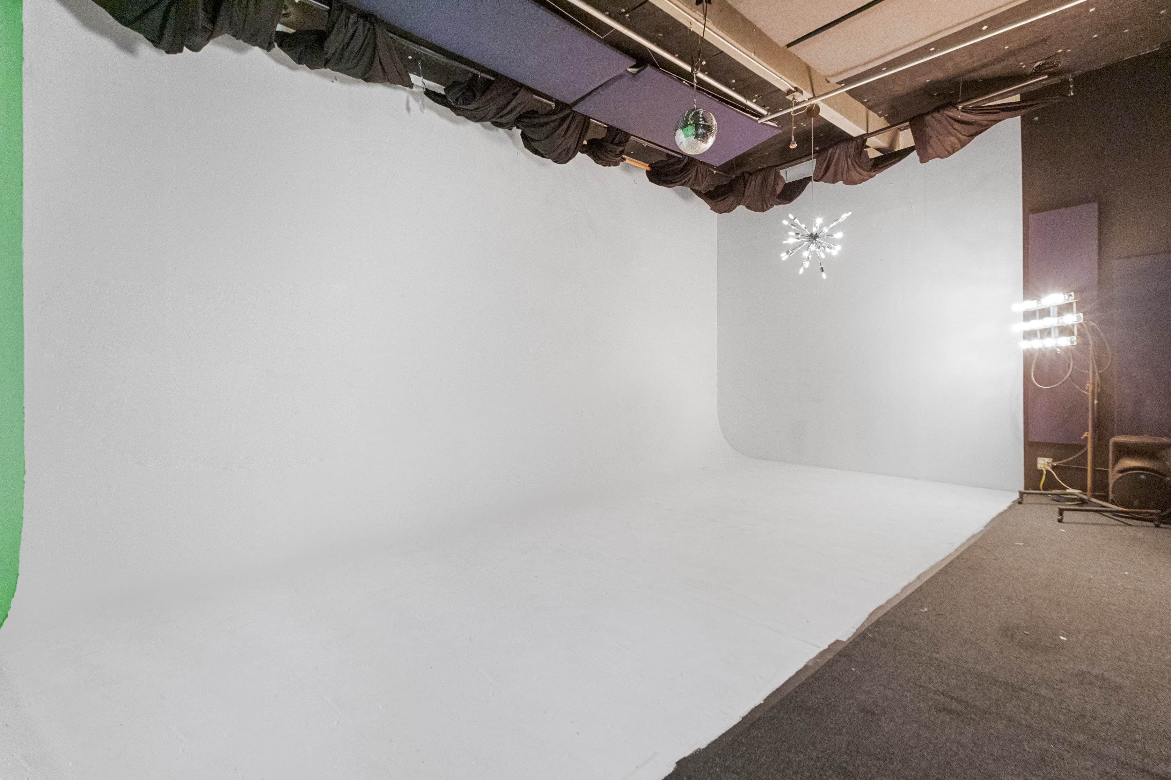 The image shows an empty studio space with a white backdrop, a green screen, and lighting equipment in the corner.