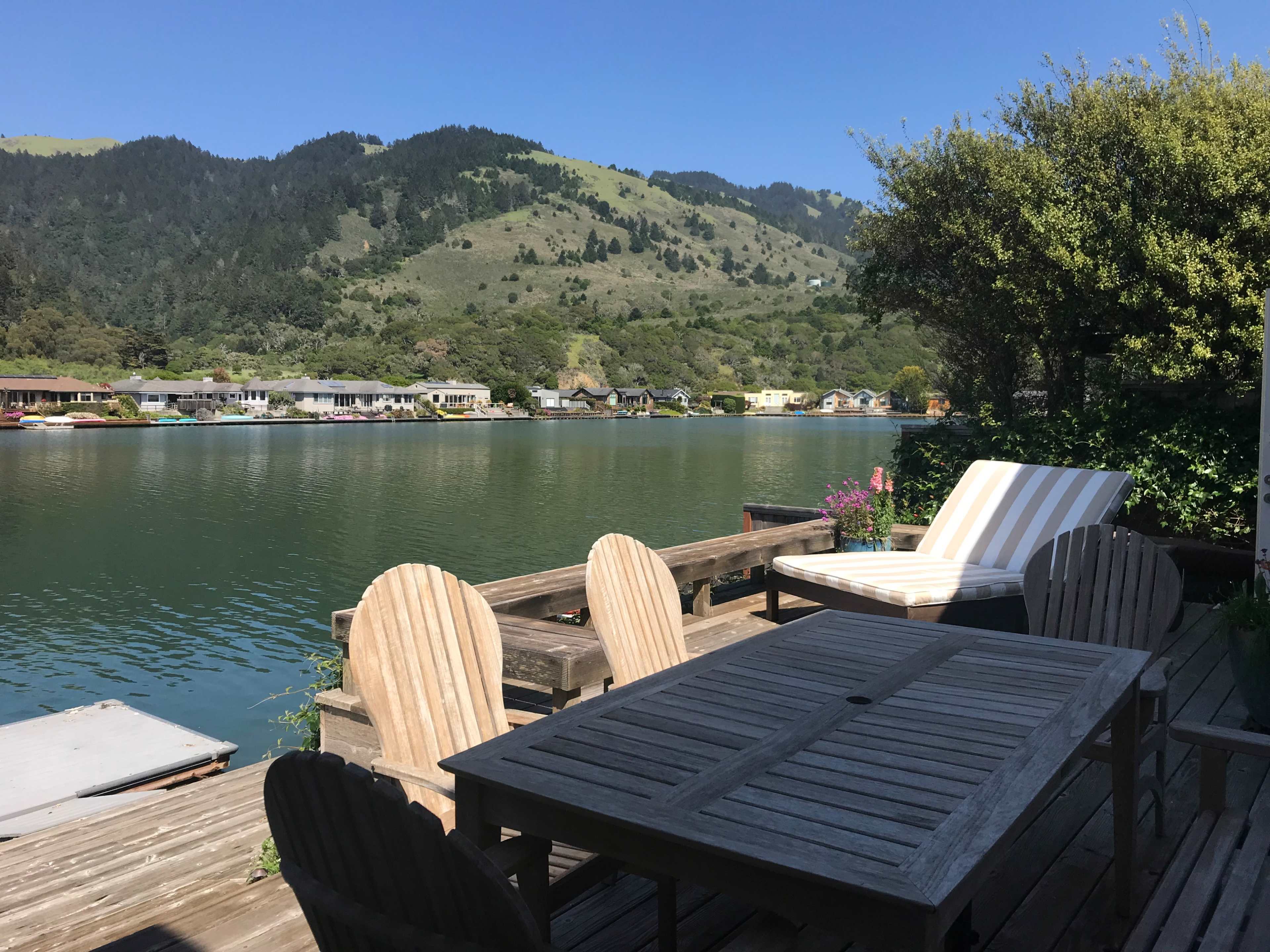 A wooden deck with patio furniture overlooks a calm waterfront and lush green hills in the background.