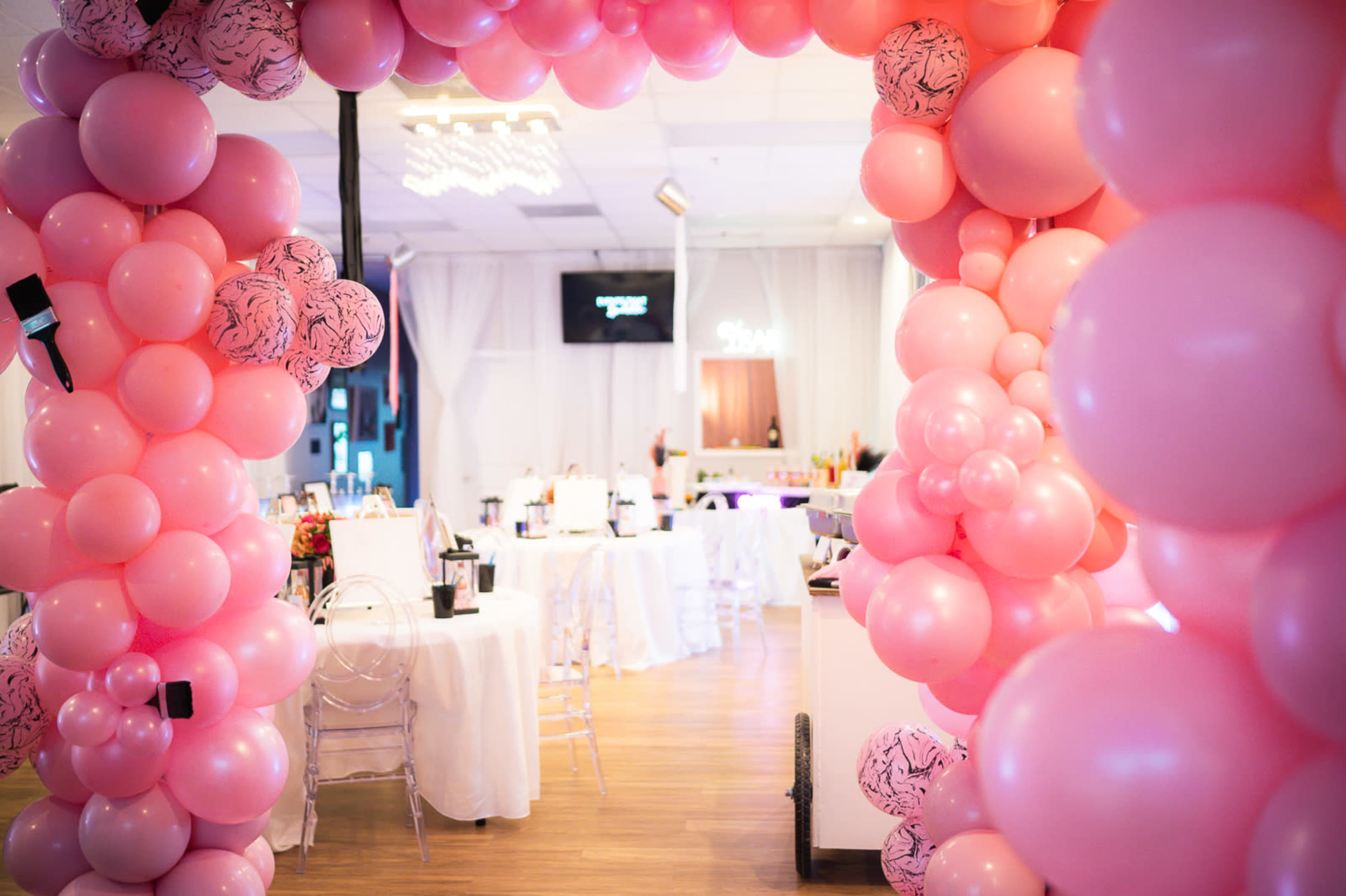 A large pink balloon arch frames an elegant event space featuring tables and decorations for a celebration.