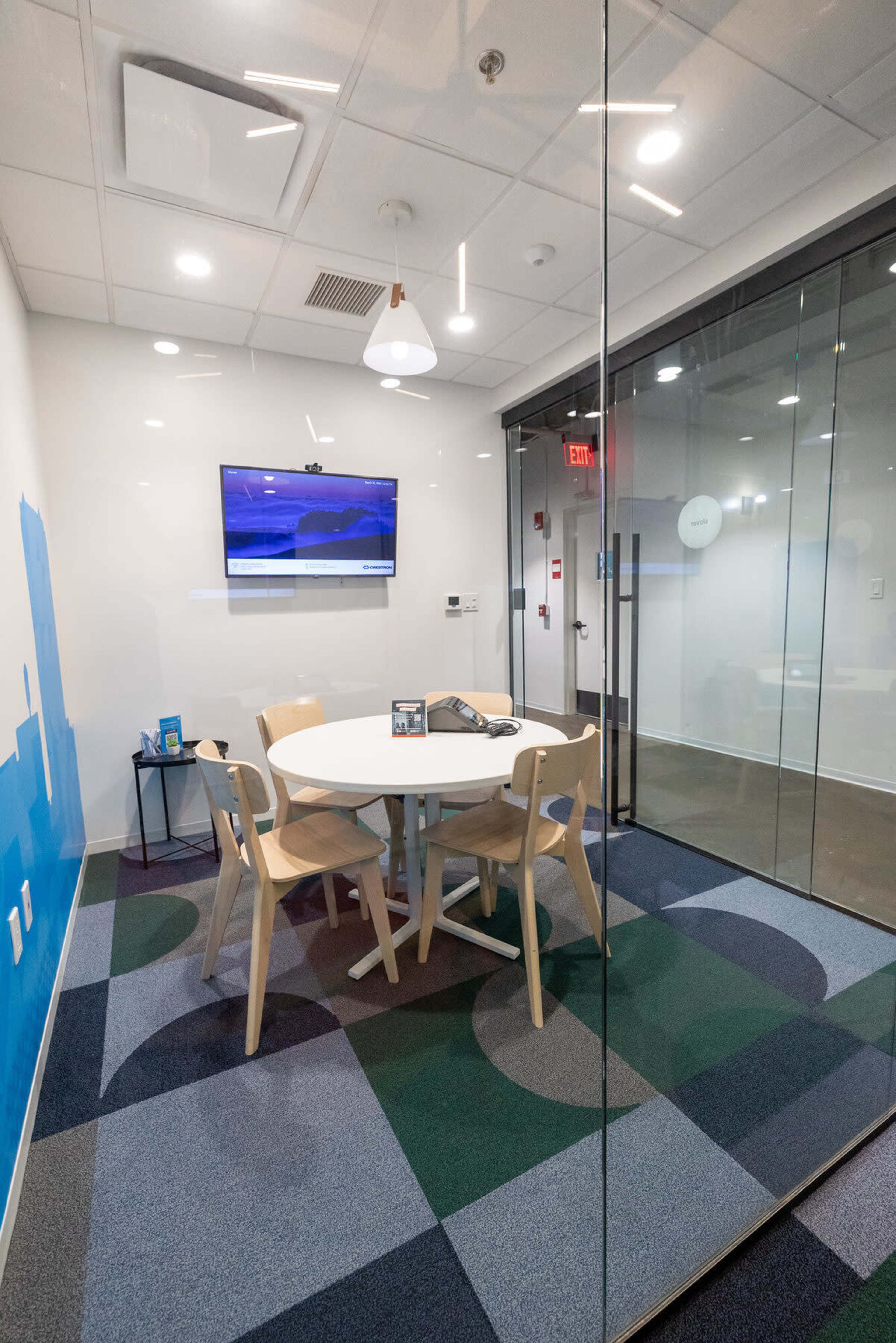 The image shows a modern conference room with a round table and four chairs, a television mounted on the wall, and large glass windows.