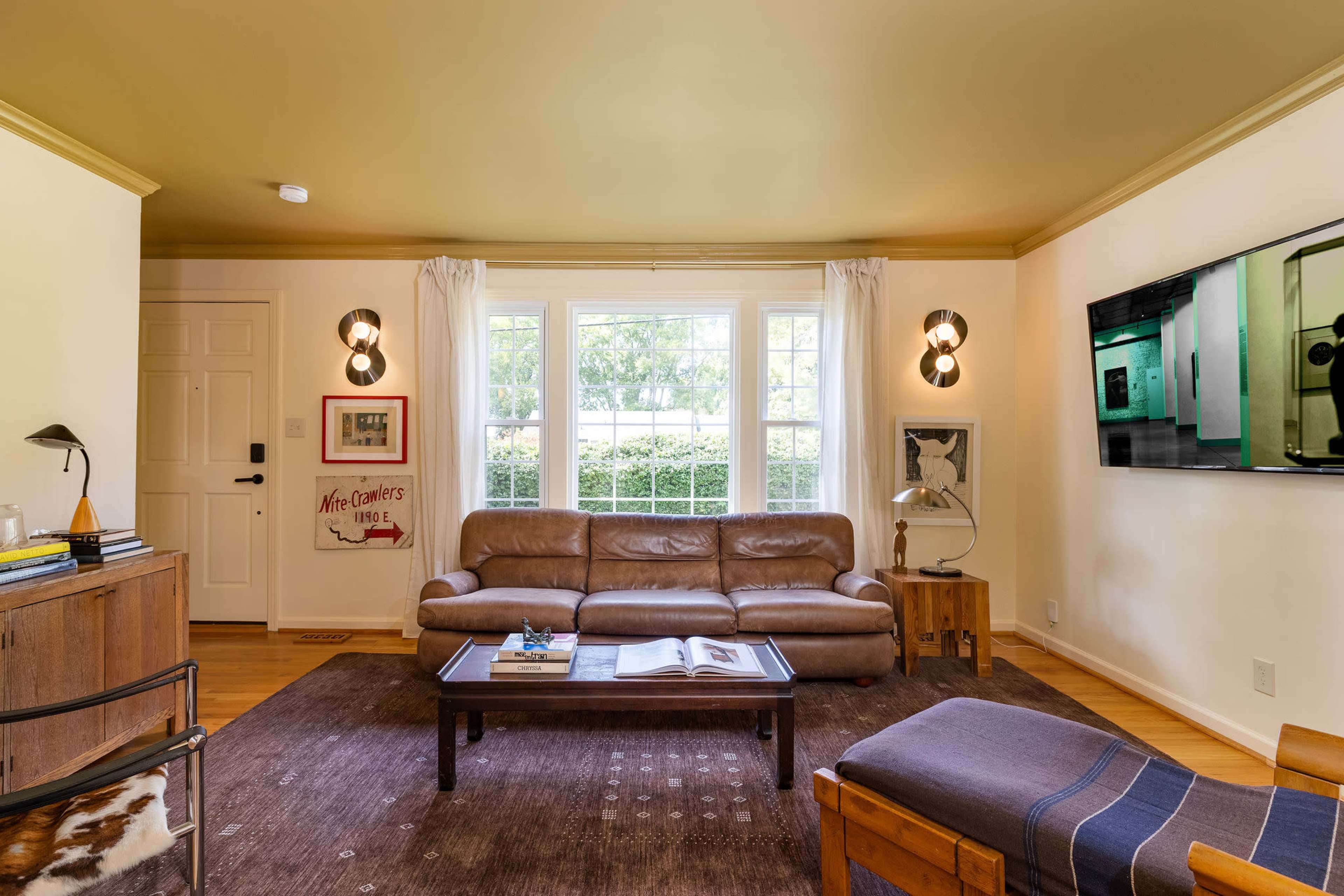 The living room features a brown leather sofa, wooden furniture, a coffee table, and large windows that overlook a garden, with a television mounted on the wall.