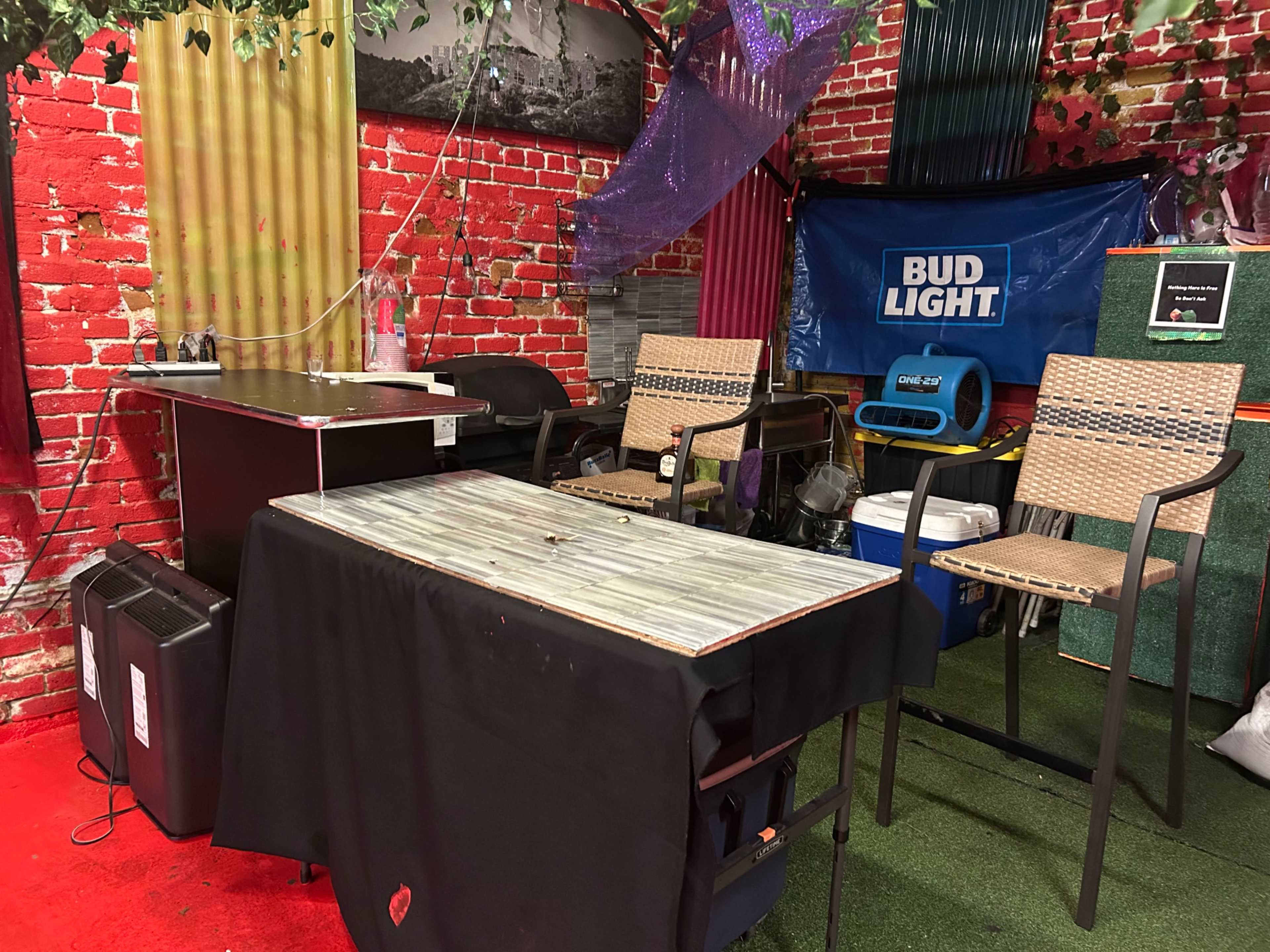The scene features a small room with a table covered by a black cloth, two tall chairs, and a backdrop of red brick walls with various decorations and a Bud Light banner.