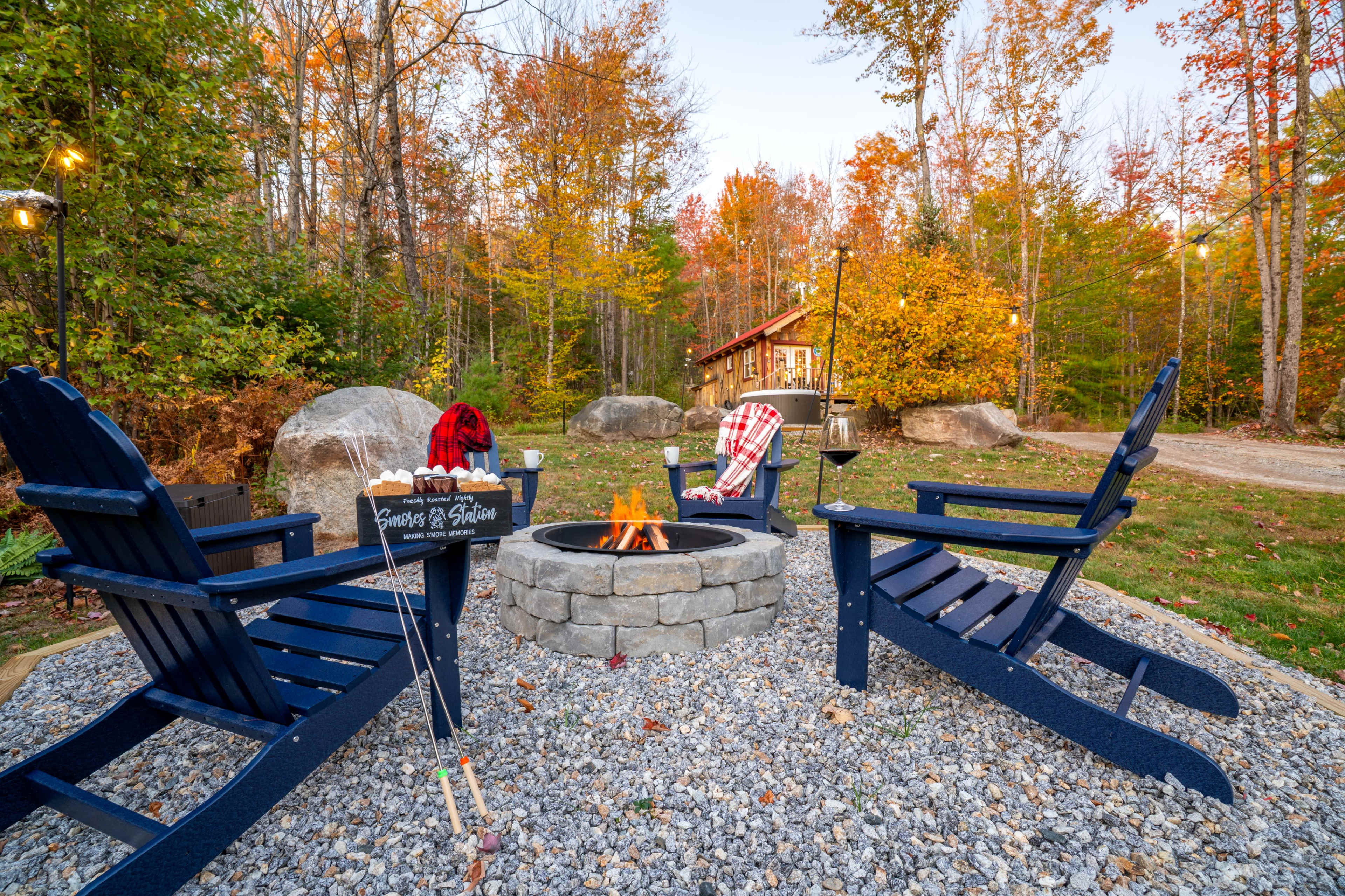 A cozy fire pit surrounded by four blue Adirondack chairs is set in a wooded area with autumn leaves and a cabin in the background.