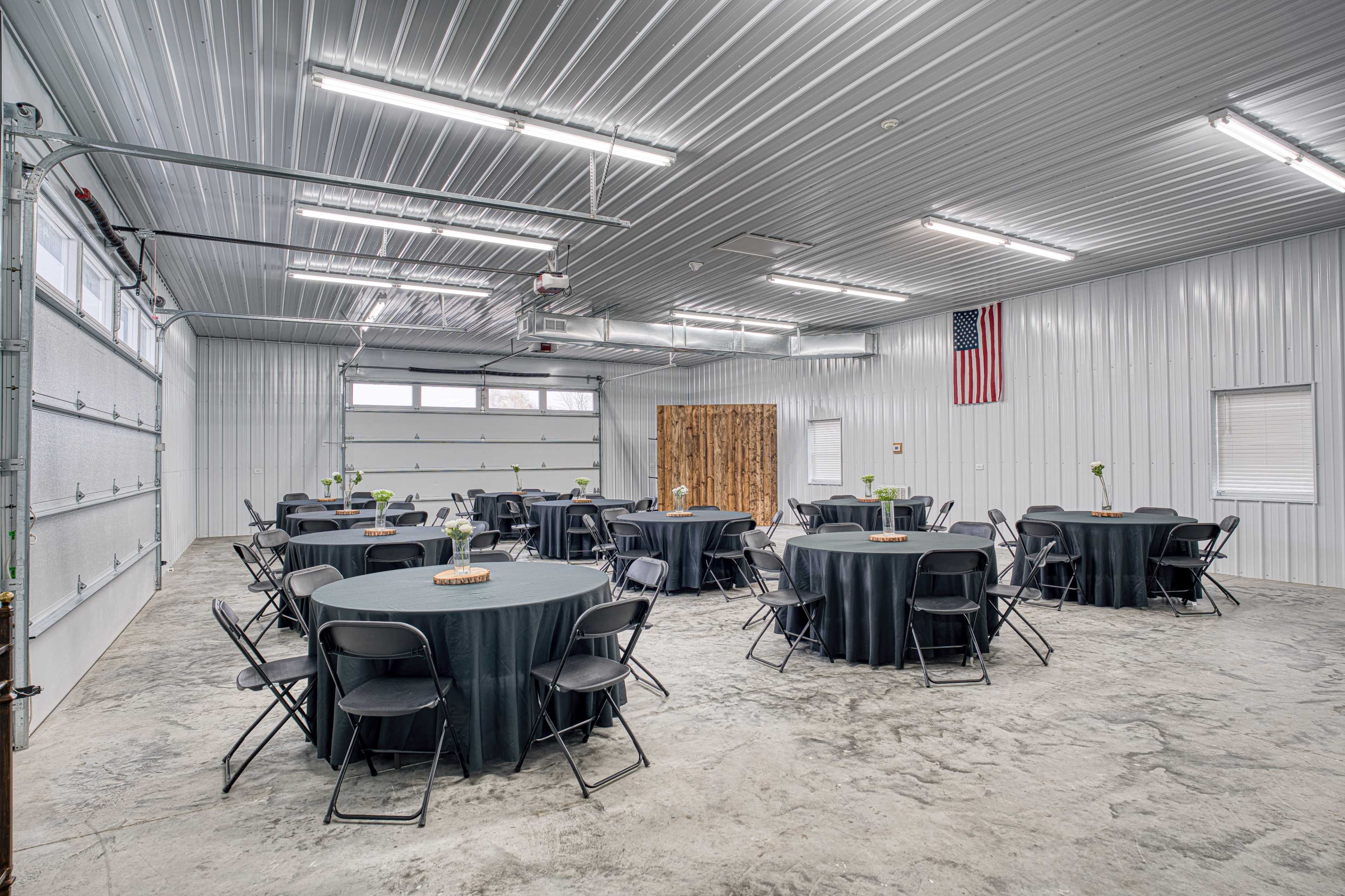 The image shows a spacious indoor venue with neatly arranged round tables covered in black tablecloths, set up for an event in a well-lit industrial-style room.